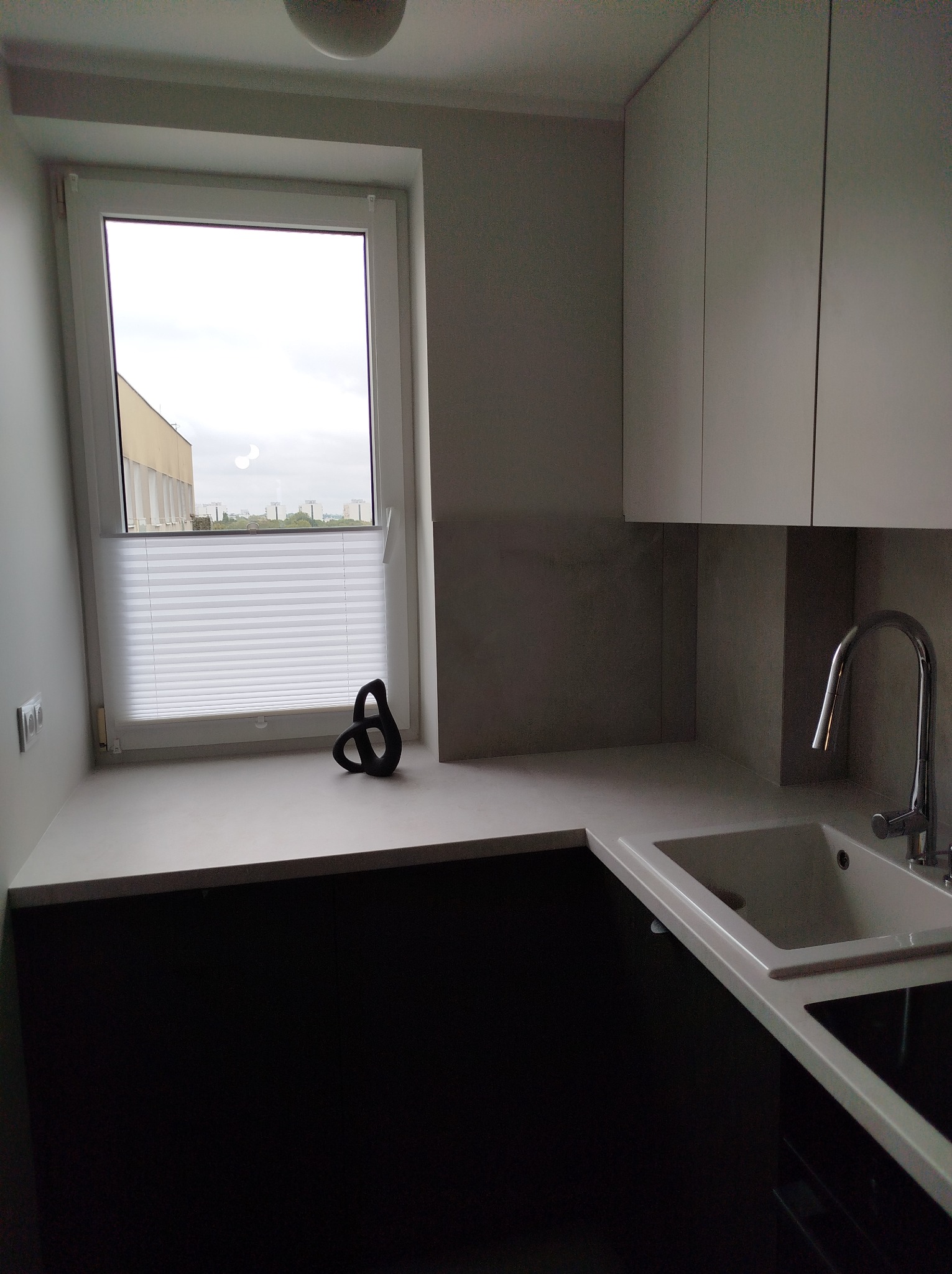 Nowoczesna kuchnia z widokiem na miasto przez okno z plisowaną roletą, czarne szafki dolne i białe górne, minimalistyczna rzeźba na blacie.