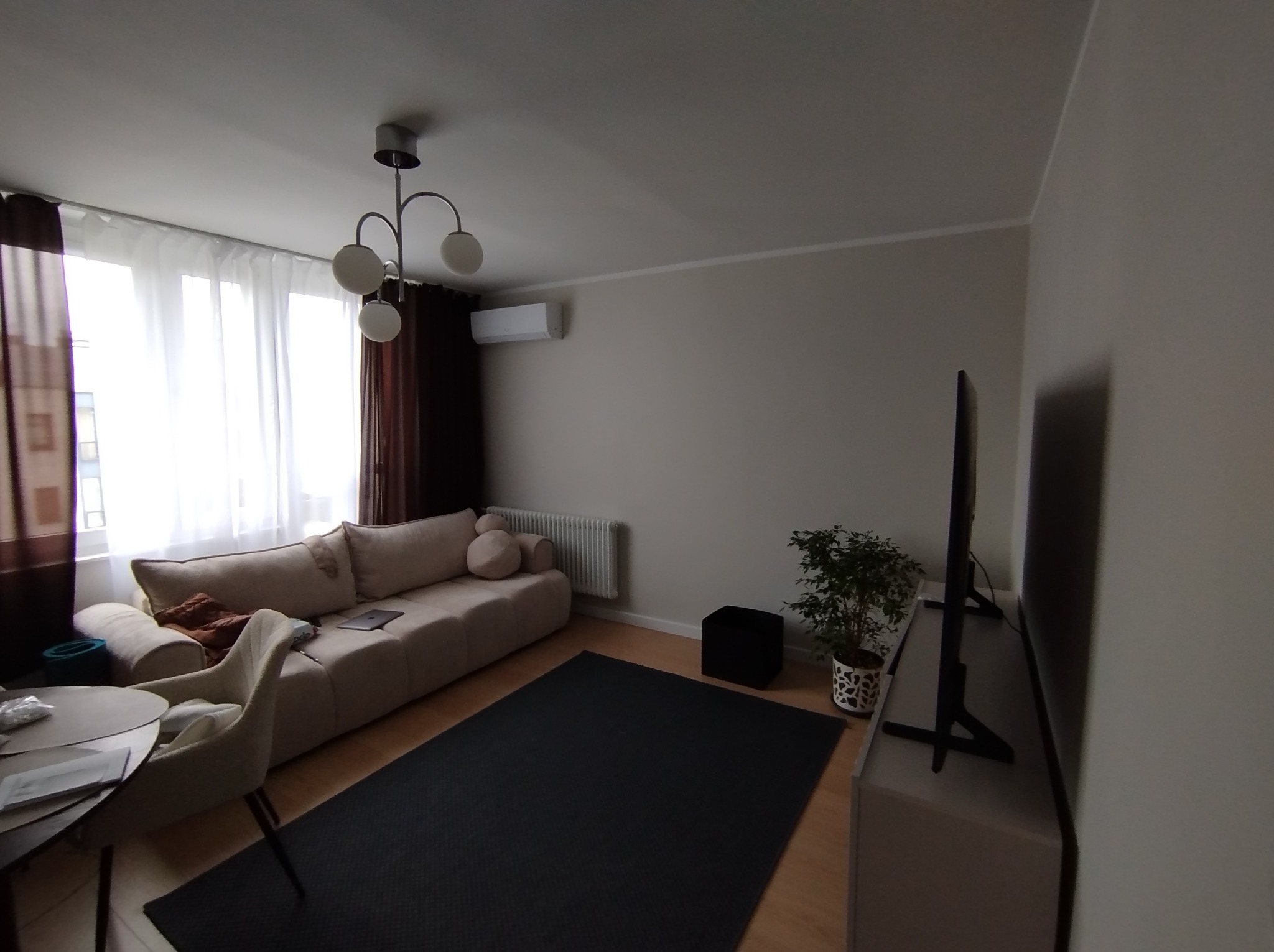 Jasne wnętrze pokoju dziennego z beżową sofą, ciemnym dywanem, telewizorem na szafce RTV i okrągłym stołem z krzesłem. Widoczny klimatyzator na ścianie i roślina doniczkowa.