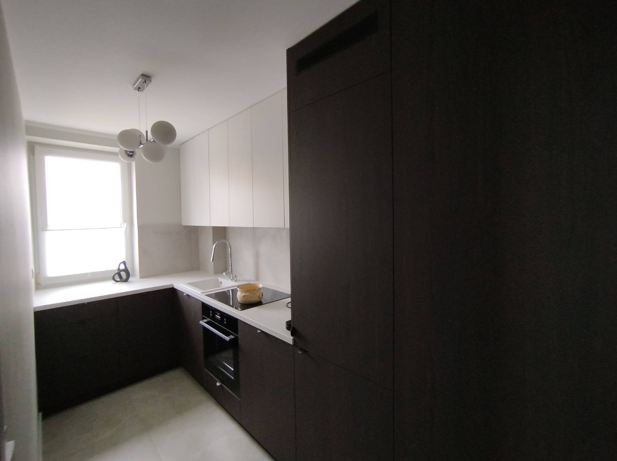 Nowoczesna kuchnia z białymi szafkami górnymi i ciemnobrązowymi dolnymi, jasny blat, okno z dekoracją, designerska lampa sufitowa, widoczny garnek na płycie grzewczej.