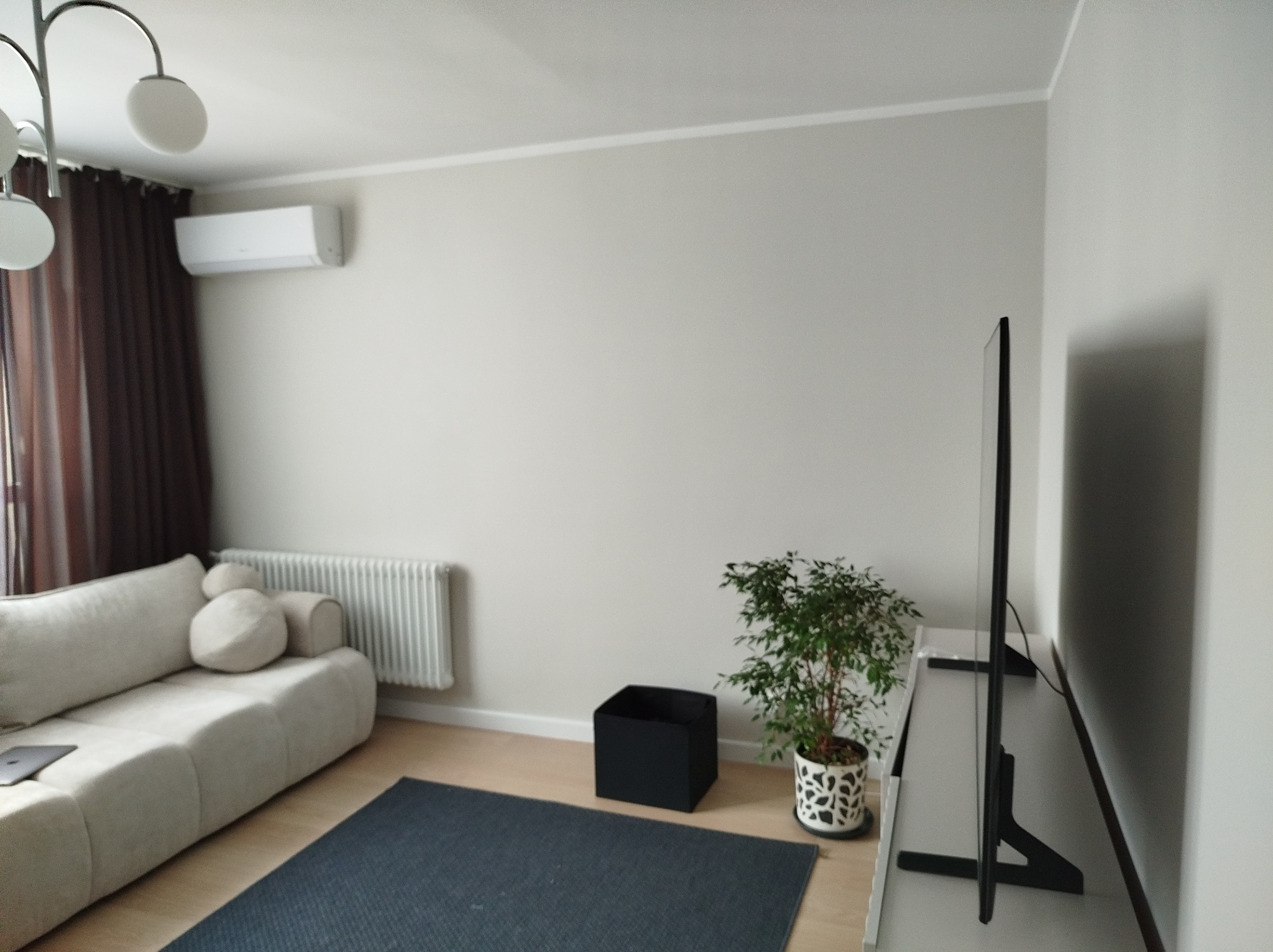 Jasne wnętrze salonu w Warszawie po odświeżeniu, z beżową sofą, ciemnym dywanem, telewizorem na niskiej szafce i rośliną doniczkową, oświetlone światłem dziennym.