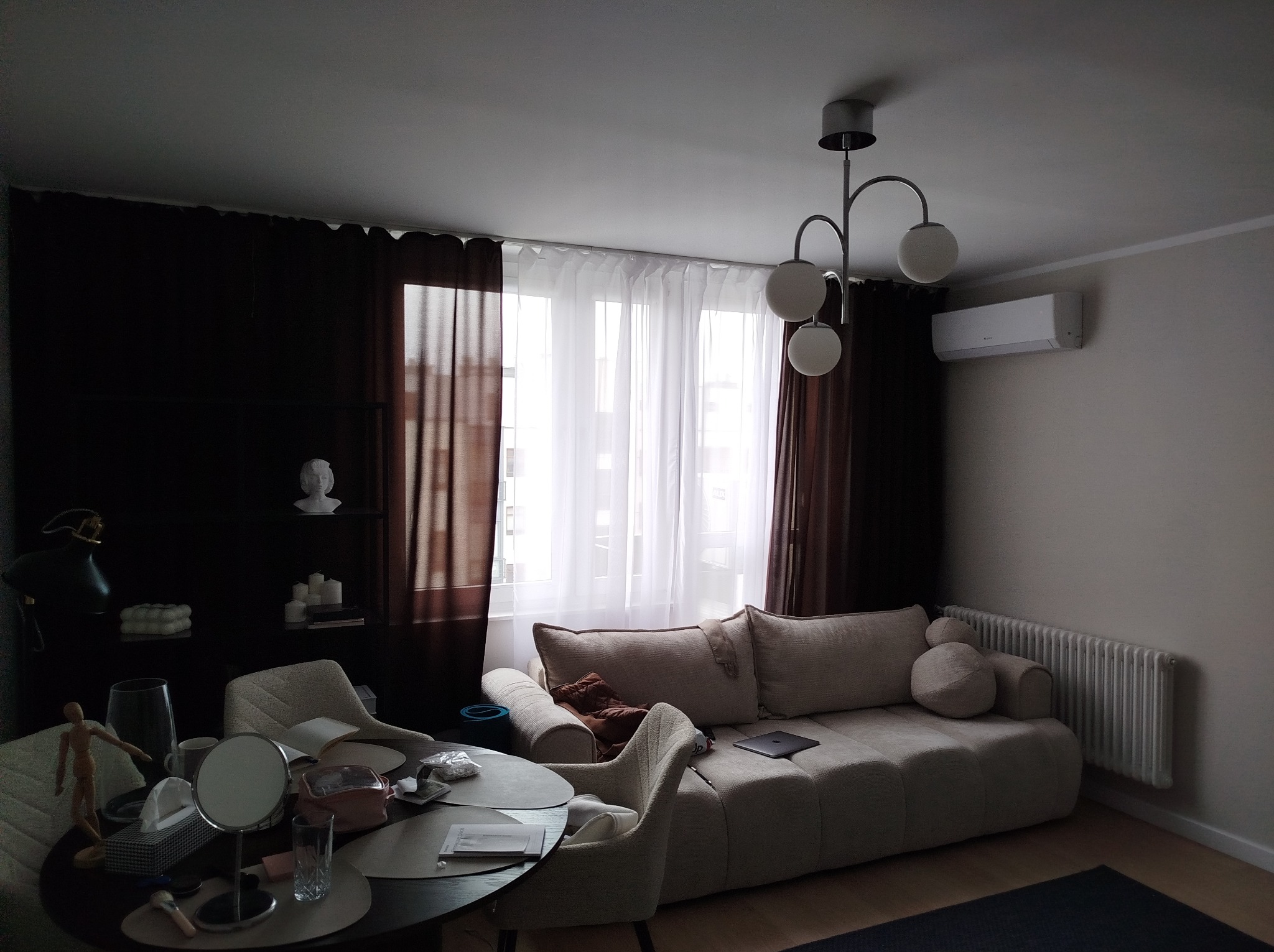 Jasny salon z beżową sofą modułową, okrągłym stolikiem kawowym z przyborami toaletowymi, ciemnymi zasłonami i nowoczesną lampą sufitową z trzema kulami.