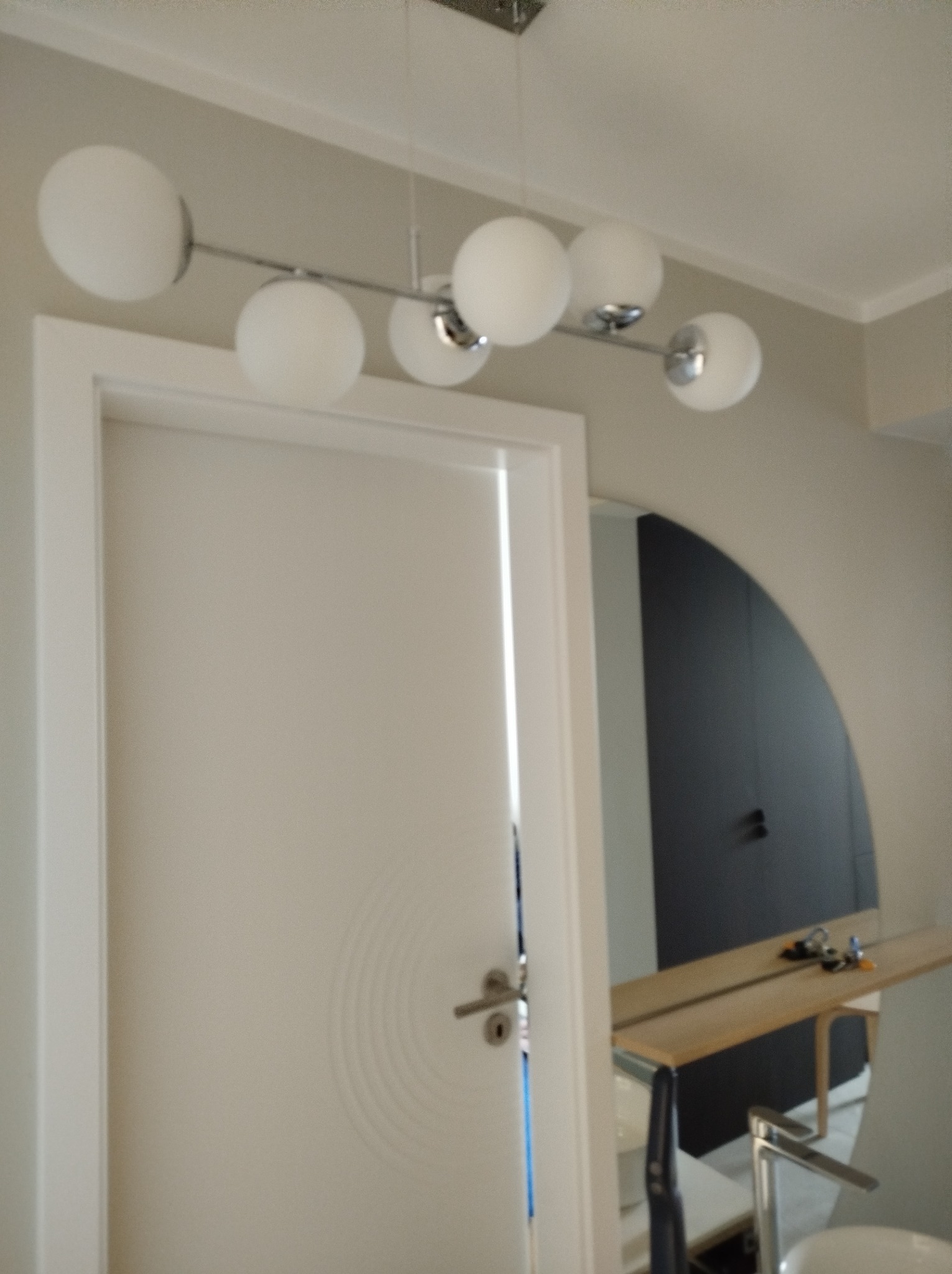 Nowoczesna lampa sufitowa z białymi kulami nad białymi drzwiami z minimalistycznym wzorem, w tle widoczne elementy łazienki z zaokrąglonym lustrem.