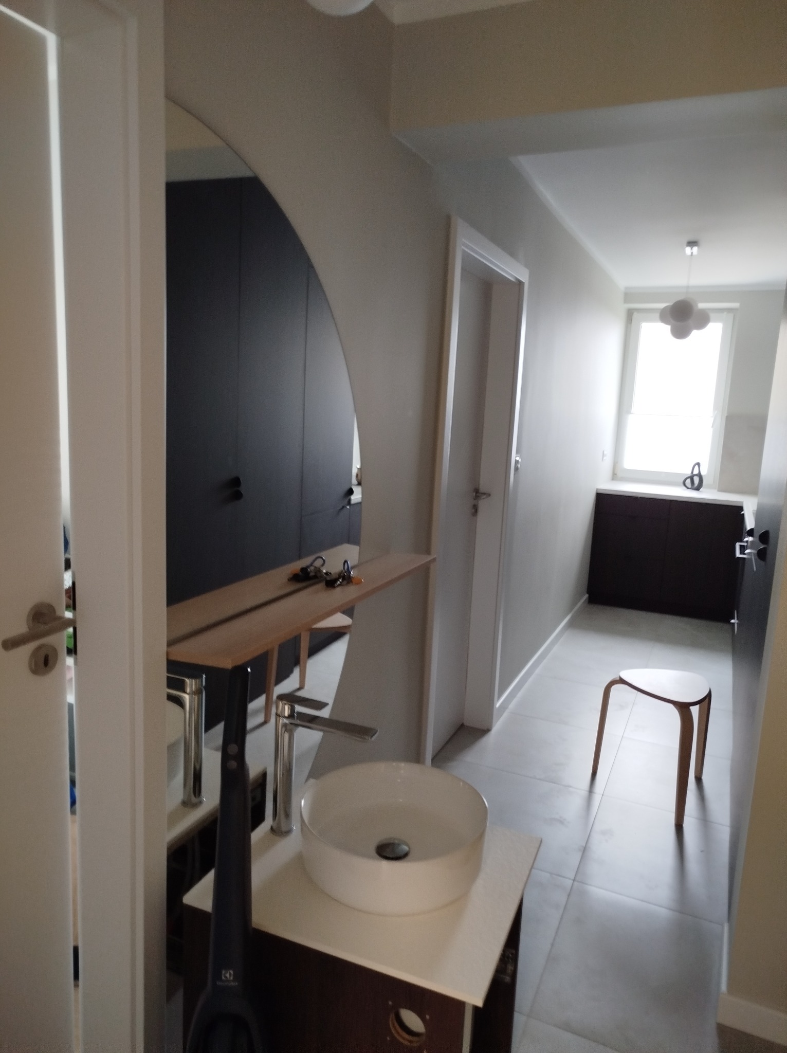 Nowoczesna łazienka z okrągłym lustrem, umywalką nablatową i ciemnymi szafkami w tle, częściowo widoczny odkurzacz pionowy.