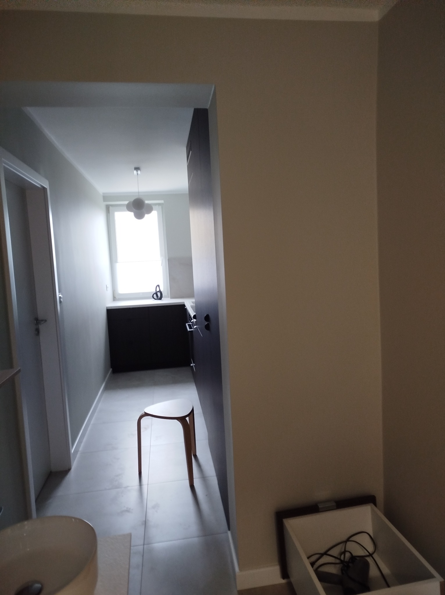 Wnętrze łazienki w trakcie prac wykończeniowych, widoczny fragment umywalki, taboret i otwarte drzwi do pomieszczenia z szafkami i oknem, a w rogu skrzynka z narzędziami.