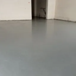 Świeżo wylana, gładka, szara posadzka w pustym pomieszczeniu z otwartym przejściem do kolejnego pokoju, gdzie widoczne są rury instalacyjne.