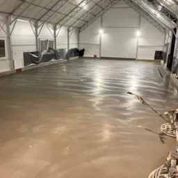 Świeżo wylana, gładka posadzka w hali przemysłowej, częściowo zabezpieczona folią, z widocznymi maszynami do zacierania i pracownikiem w tle po prawej stronie.