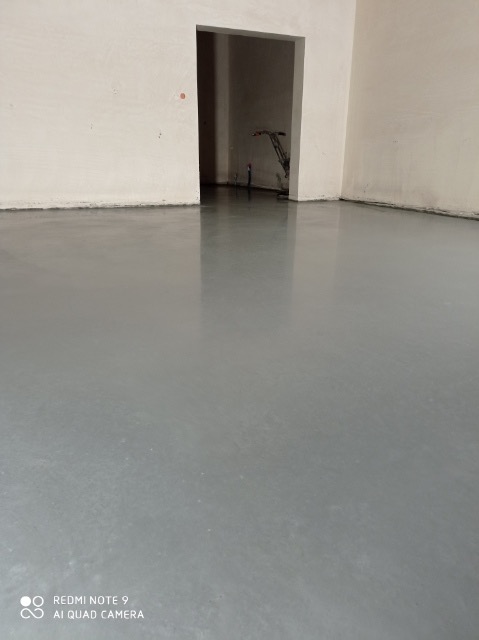 Świeżo wylana, gładka, szara posadzka w pustym pomieszczeniu z otwartym przejściem do kolejnego pokoju, gdzie widoczne są rury instalacyjne.