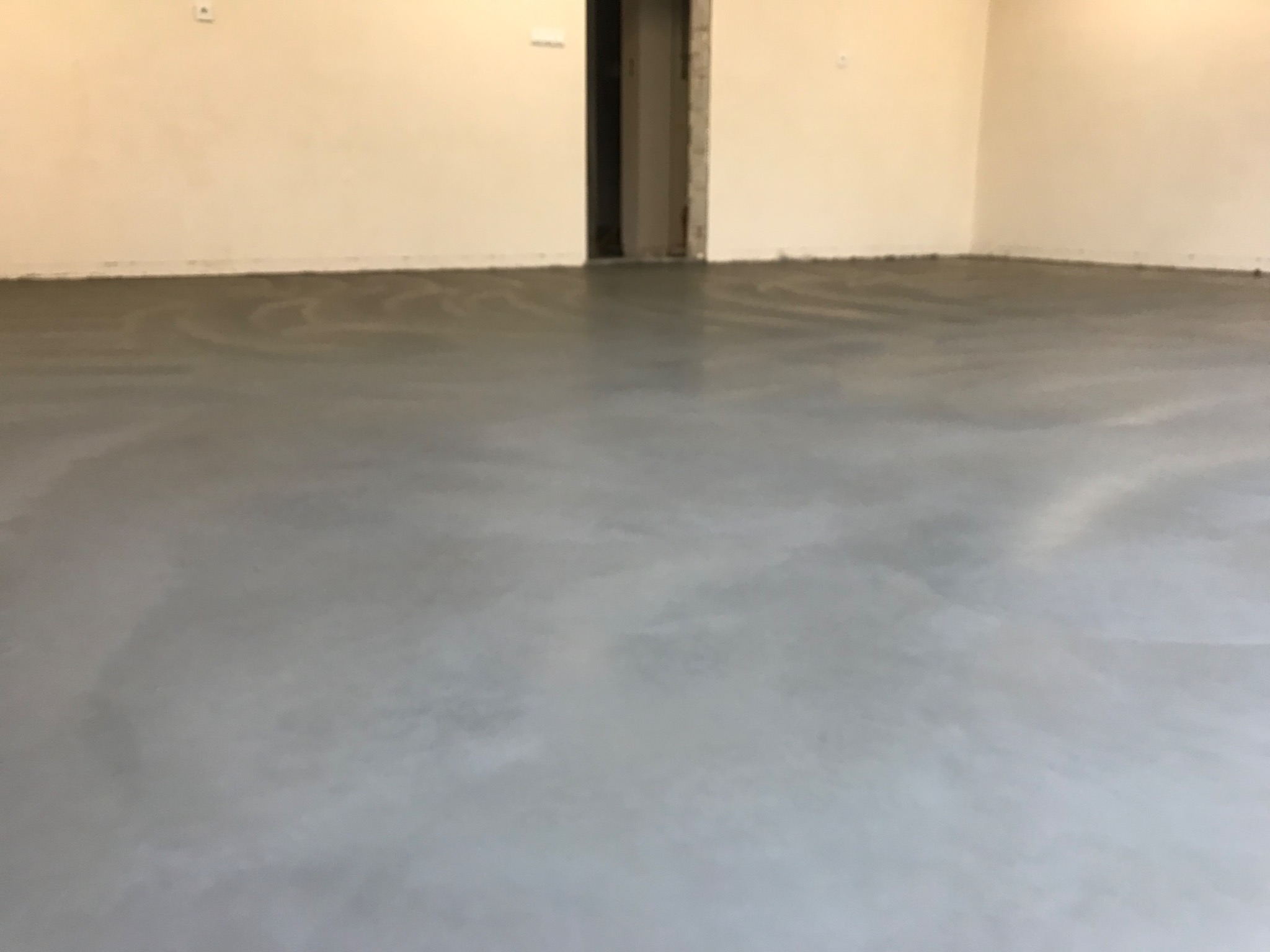 Świeżo wylana, gładka, szara posadzka anhydrytowa w pustym pomieszczeniu z widocznym wejściem bez drzwi.