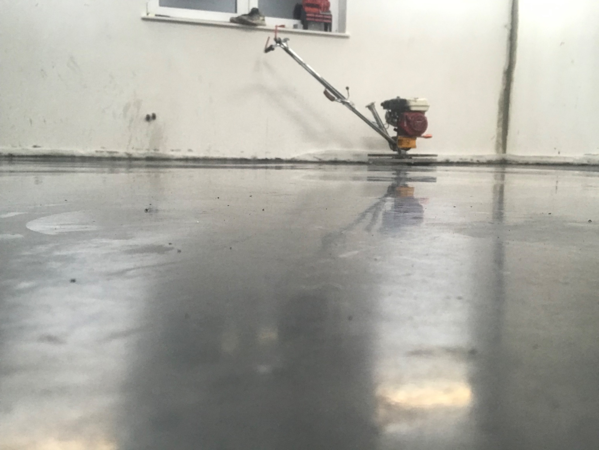 Świeżo wylana, mokra posadzka zacierana mechaniczną zacieraczką. W tle widoczne buty i narzędzia na parapecie okna.