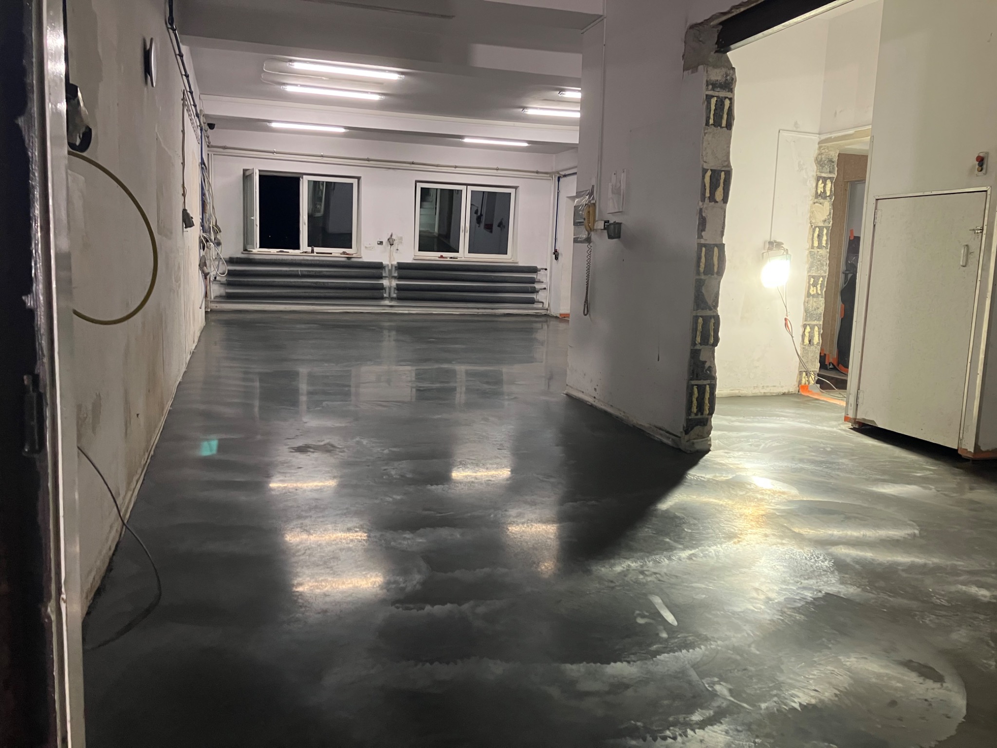 Świeżo wylana, błyszcząca, szara posadzka w dużym, pustym pomieszczeniu przemysłowym z widocznymi śladami po procesie wylewania, oświetlona przez lampy sufitowe i lampę roboczą.