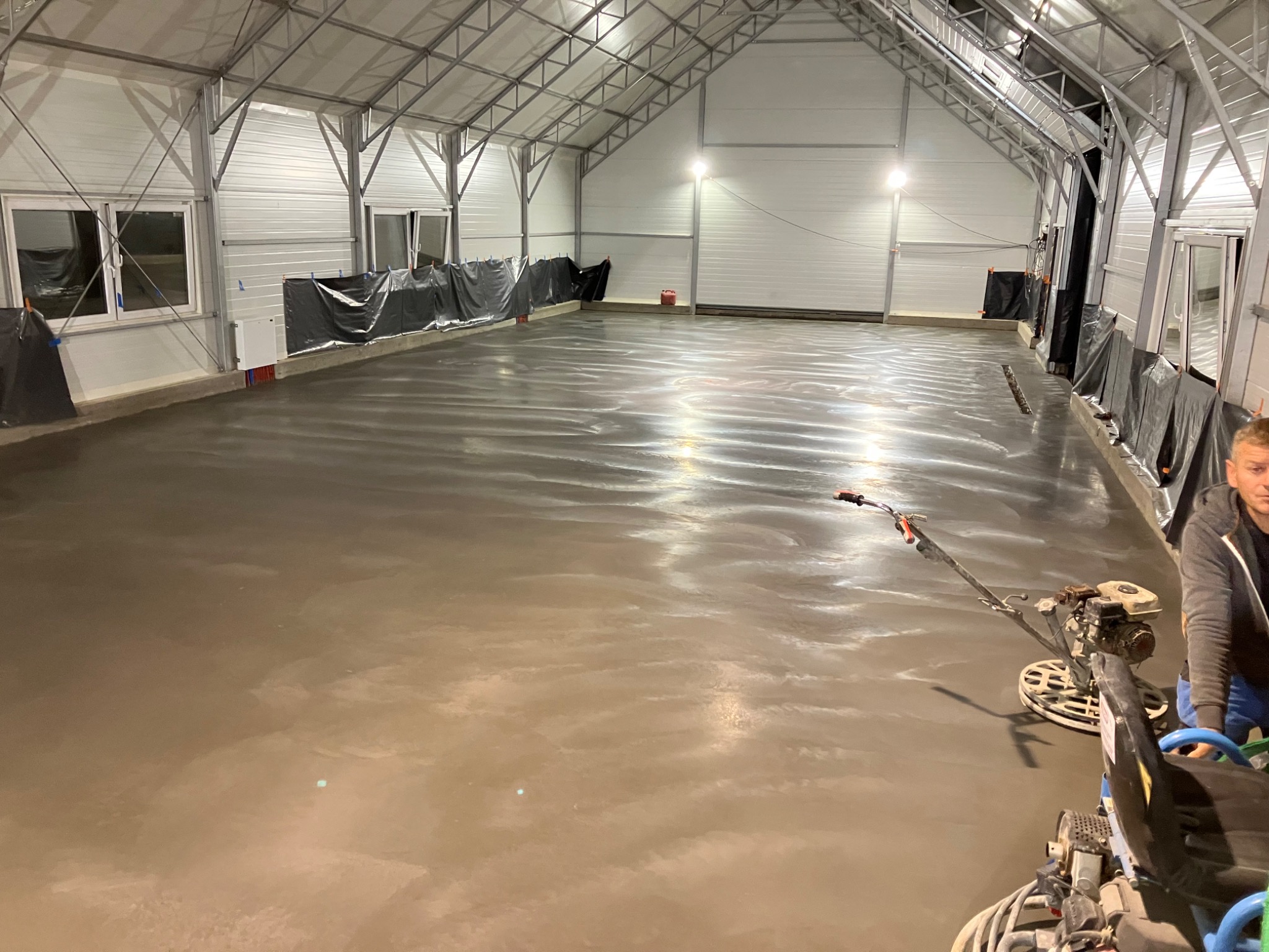 Świeżo wylana, gładka posadzka w hali przemysłowej, częściowo zabezpieczona folią, z widocznymi maszynami do zacierania i pracownikiem w tle po prawej stronie.