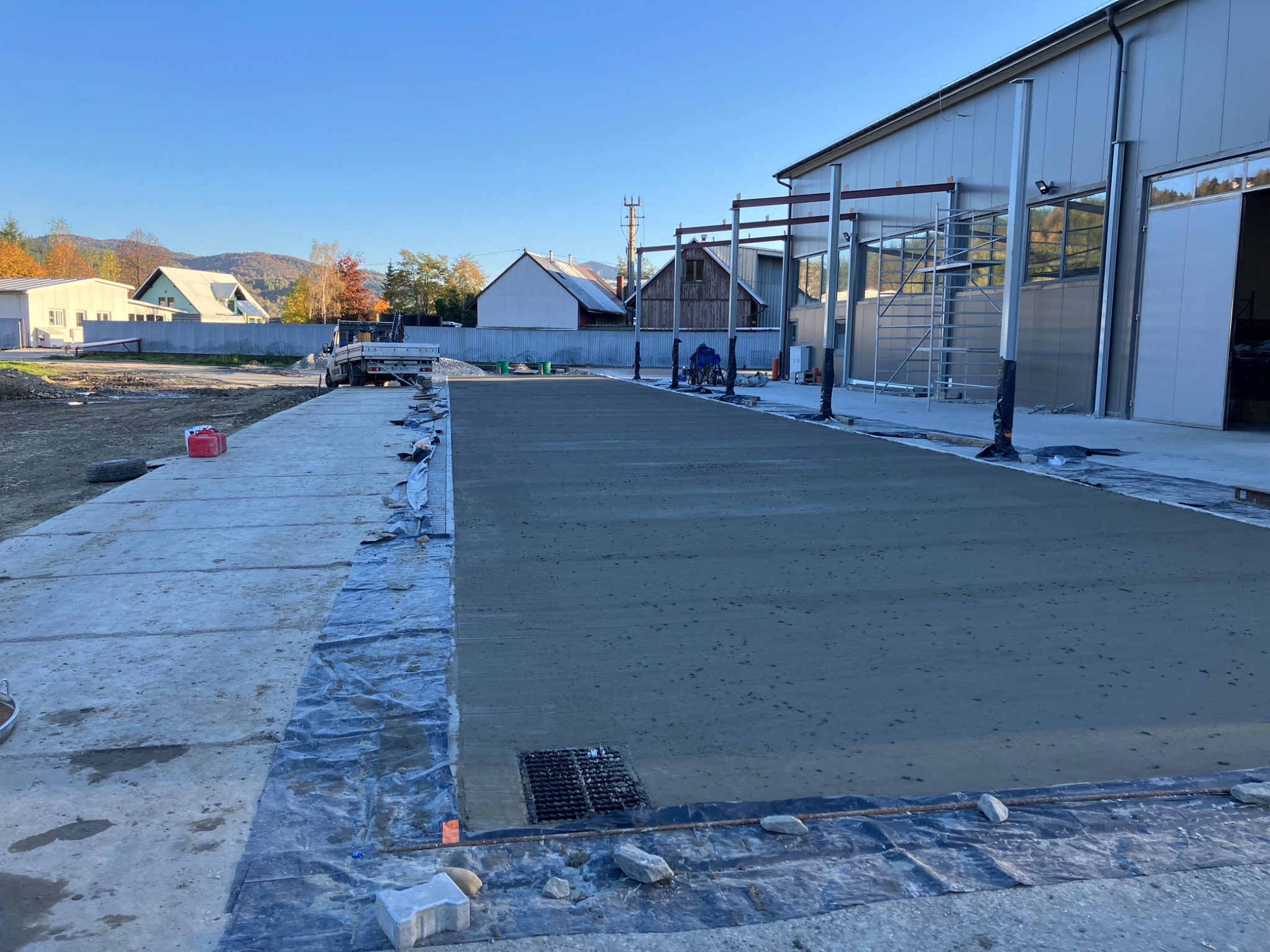 Świeżo wylana, gładka posadzka na zewnątrz budynku przemysłowego, z widocznym odpływem liniowym i przygotowanym podłożem, w tle krajobraz zabudowań i wzgórz pod błękitnym niebem.