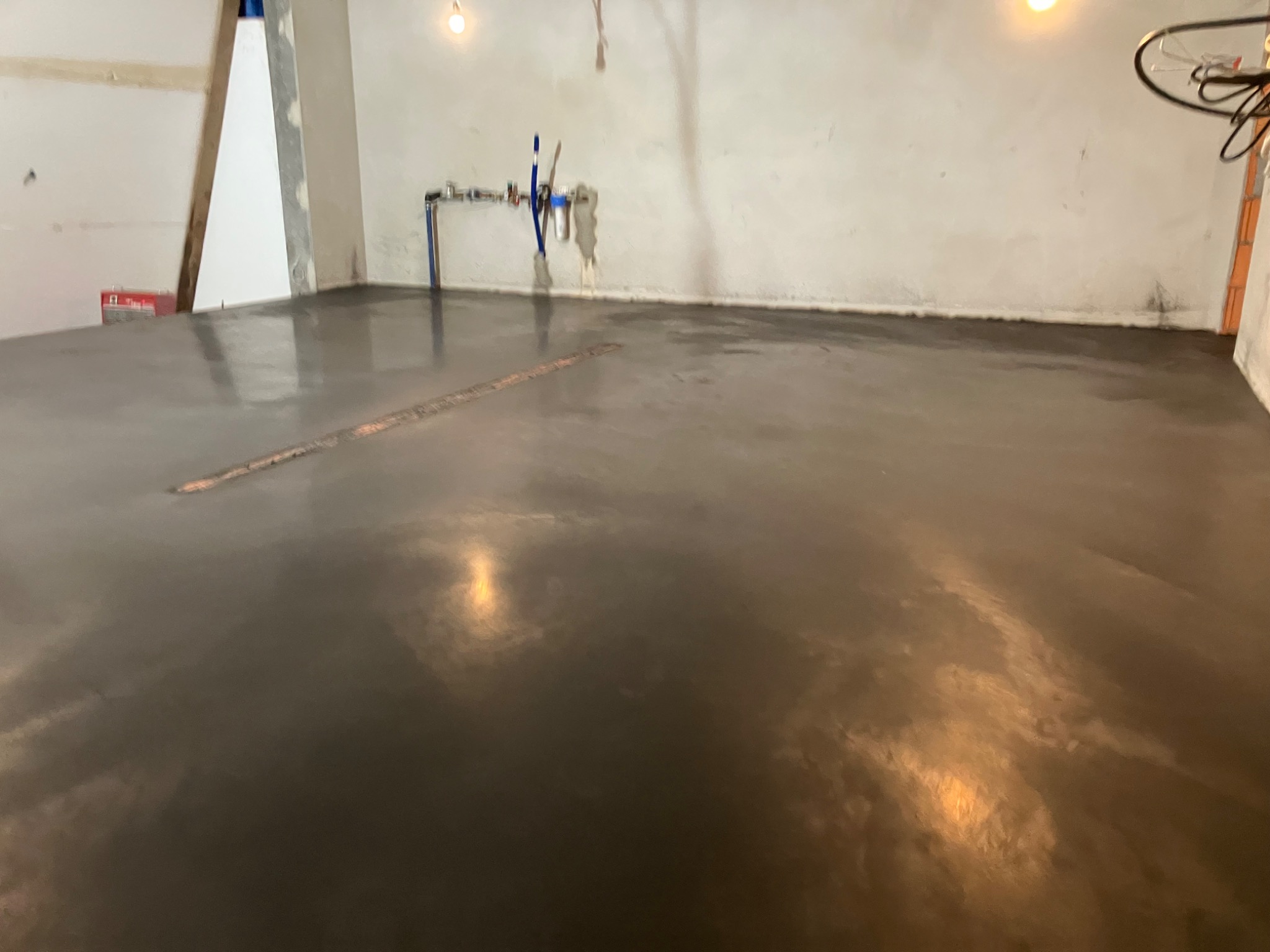 Świeżo wylana, gładka, szara posadzka w surowym pomieszczeniu z instalacją wodną na ścianie i widocznymi śladami po narzędziach do rozprowadzania wylewki.