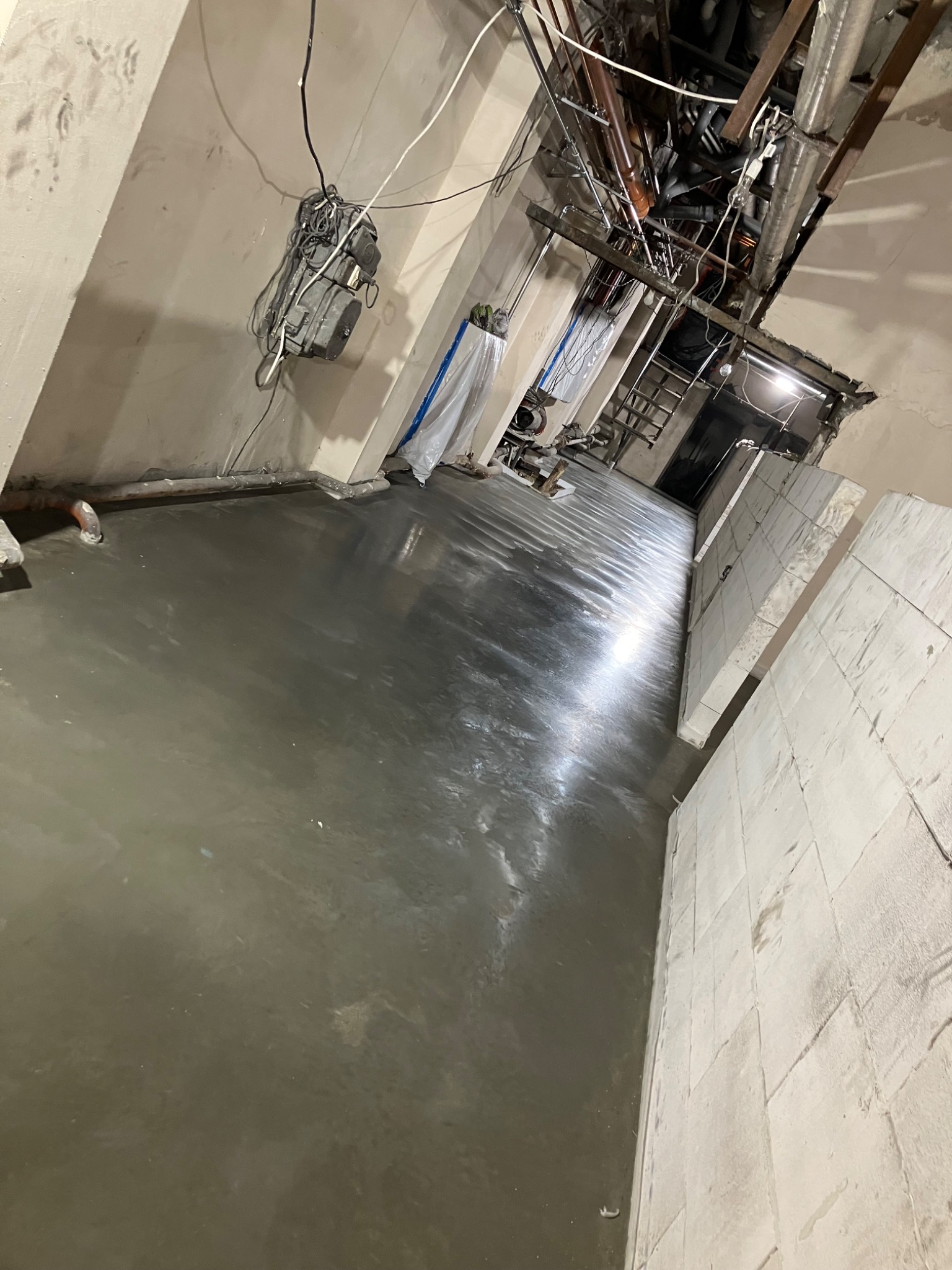 Świeżo wylana, mokra, szara posadzka w pomieszczeniu w trakcie remontu, widoczne instalacje pod sufitem i fragmenty ścian.