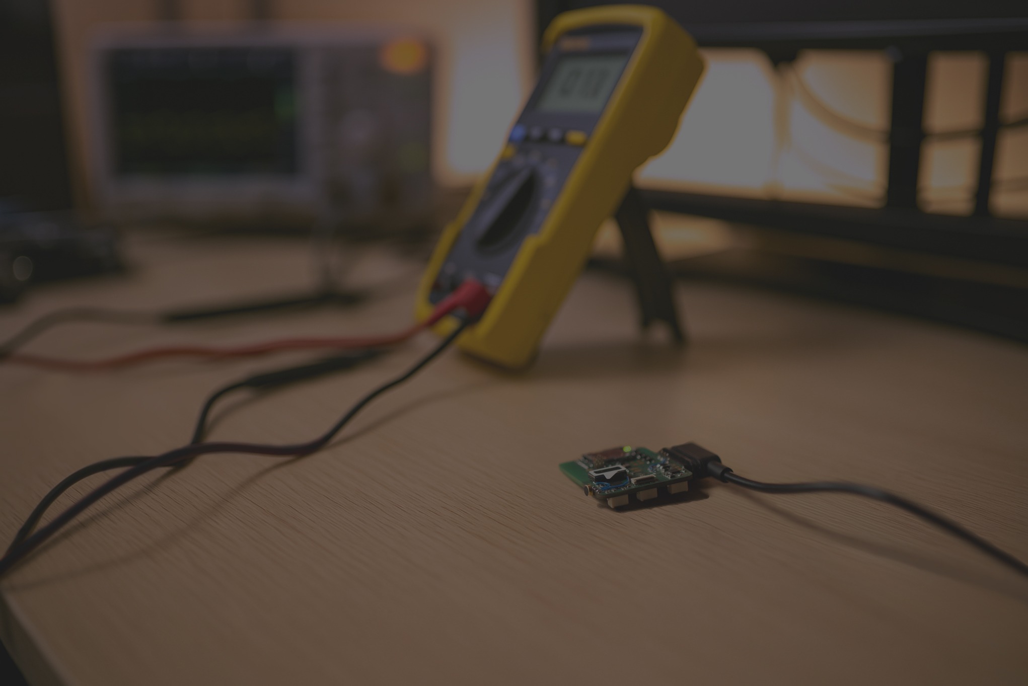 Multimetr podłączony do płytki drukowanej na drewnianym biurku, w tle oscyloskop i monitor. Sprzęt elektroniczny używany do testowania i debugowania oprogramowania.