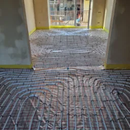 Rozłożone rury ogrzewania podłogowego na folii izolacyjnej z rastrem, widok na otwarte drzwi balkonowe i budynek gospodarczy w tle.