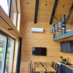 Wnętrze domu z drewnianą boazerią: widoczny biały klimatyzator ścienny, stół z żywicą epoksydową, ciemnoniebieskie szafki kuchenne i nowoczesne lampy wiszące.