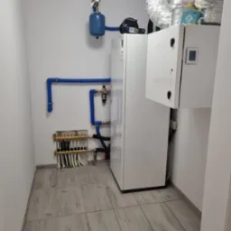 Pomieszczenie techniczne z białą jednostką rekuperacyjną Komfovent, niebieskim zbiornikiem wyrównawczym i rozdzielaczem ogrzewania podłogowego z licznymi białymi rurkami.