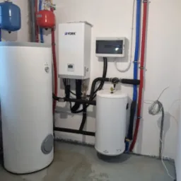 Instalacja kotłowni z białym zasobnikiem ciepłej wody, zbiornikiem wyrównawczym, rurami w kolorach niebieskim i czerwonym oraz skrzynką elektryczną na ścianie.