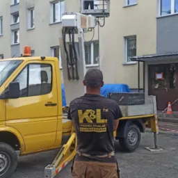 Pracownik w podnośniku koszowym czyści elewację bloku, widok zza pleców drugiego pracownika z logiem firmy na koszulce.