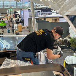 K&L profesjonalne czyszczenie - Pracownik w czarnej koszulce z logo firmy czyści wnętrze luksusowego jachtu na hali wystawienniczej, używając szmatki i rękawic ochronnych.