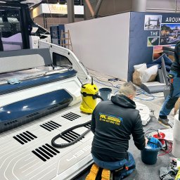 K&L profesjonalne czyszczenie - Dwaj mężczyźni w roboczych ubraniach czyszczą wnętrze luksusowej łodzi na wystawie, używając odkurzacza przemysłowego i środków czyszczących, widoczny żółty odkurzacz i butelka środka...