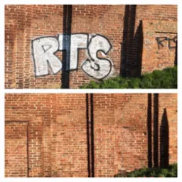 Fragment ceglanej ściany z graffiti w kolorze białym i czarnym, częściowo zasłoniętej przez cień, z zieloną trawą u podstawy.