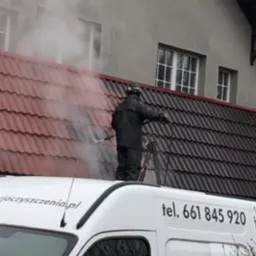 Pracownik w kombinezonie ochronnym, stojący na drabinie opierającej się o dach, czyści dachówkę za pomocą myjki ciśnieniowej; widoczny fragment białego samochodu firmowego z numerem telefonu.