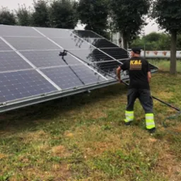 Pracownik w czarnej koszulce z logo K&L czyści panele słoneczne długą szczotką z wodą na tle zielonej trawy i drzew