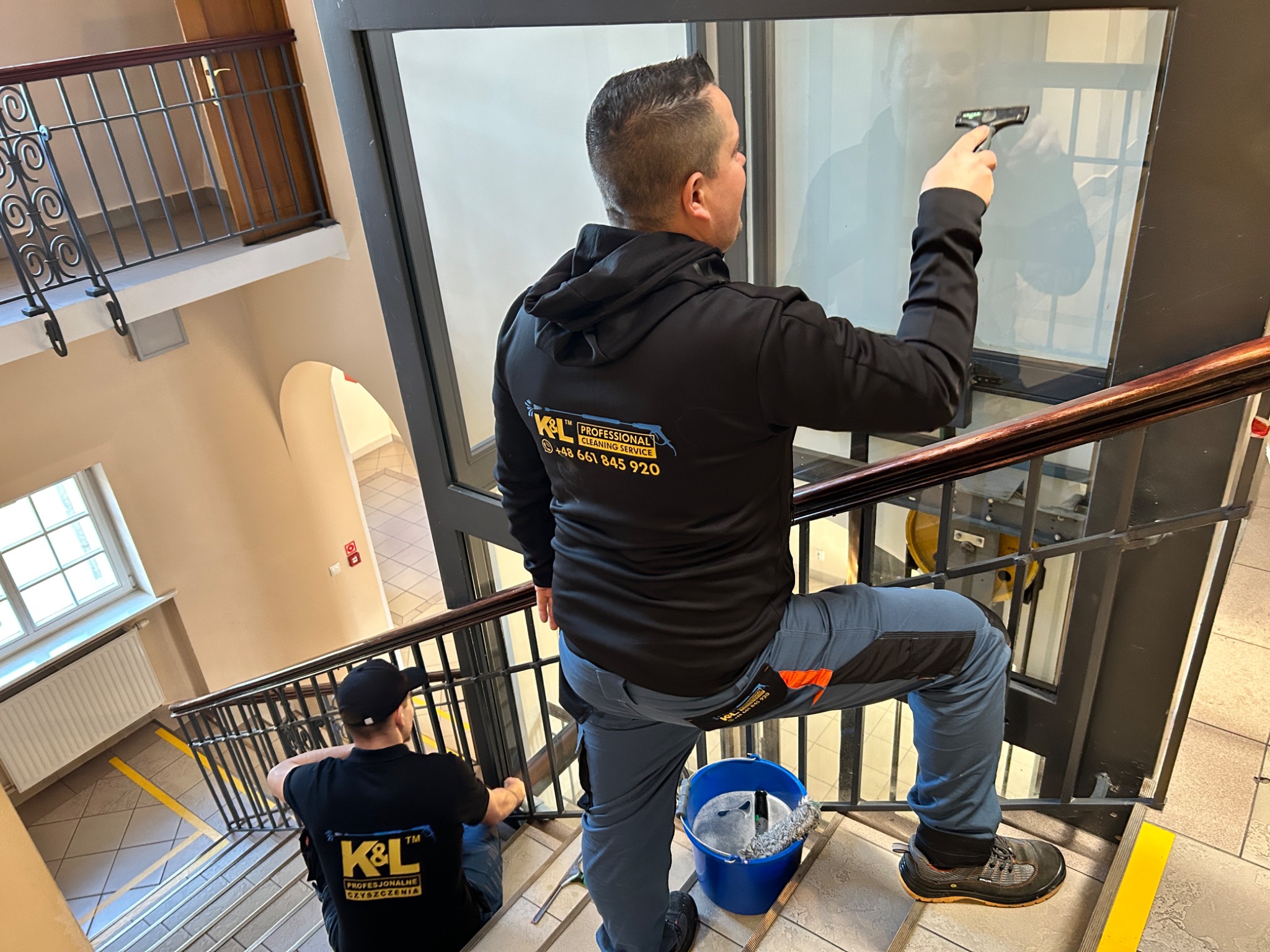 Pracownik w czarnej bluzie z logo firmy czyści szklaną ścianę na klatce schodowej za pomocą ściągaczki, drugi pracownik w czarnej koszulce z logo firmy siedzi na schodach, obok wiadro z wodą...