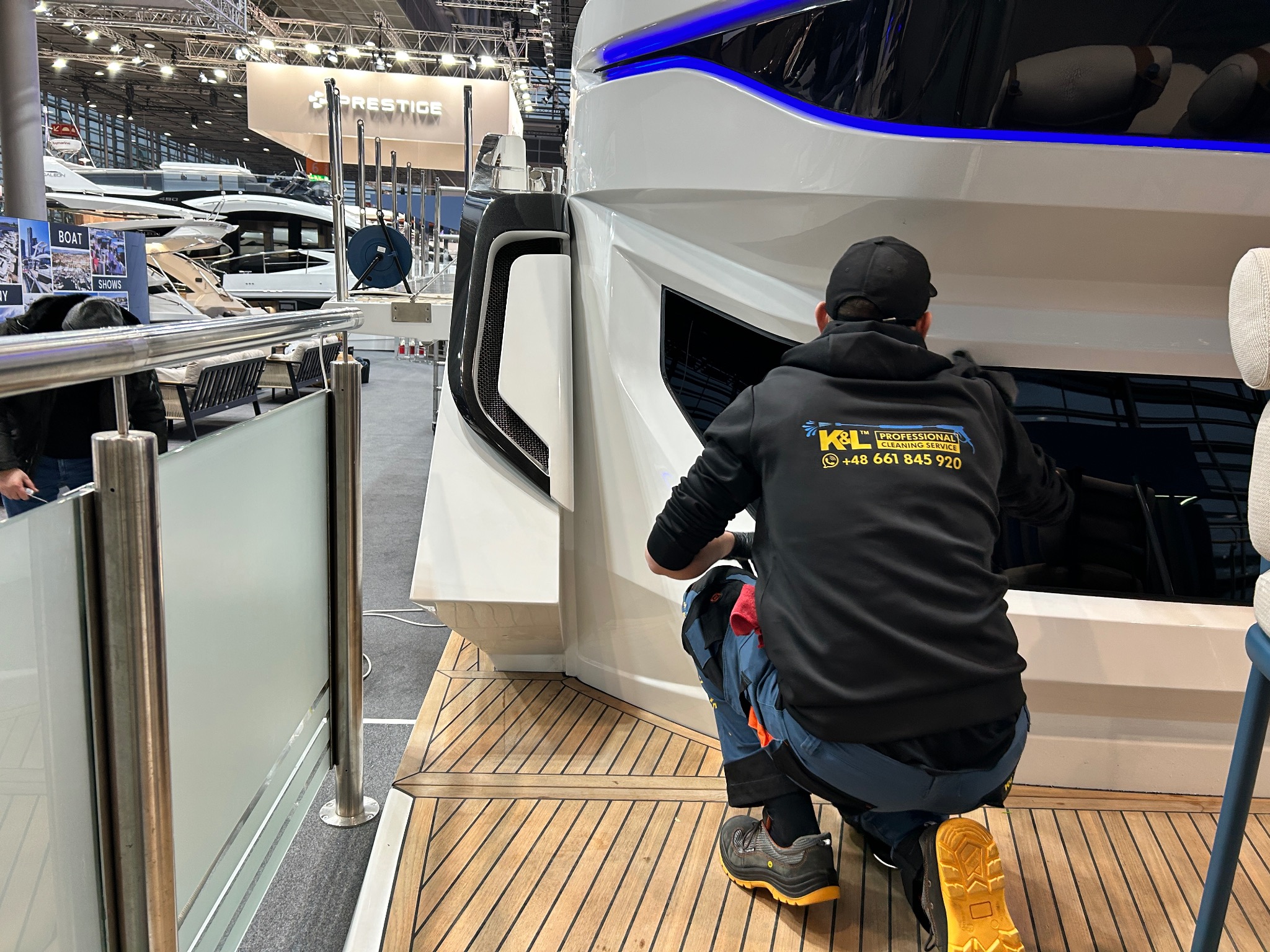 Pracownik firmy sprzątającej w czarnej bluzie z logo, klęczący na drewnianym pokładzie jachtu, czyści okno. W tle inne jachty na wystawie.