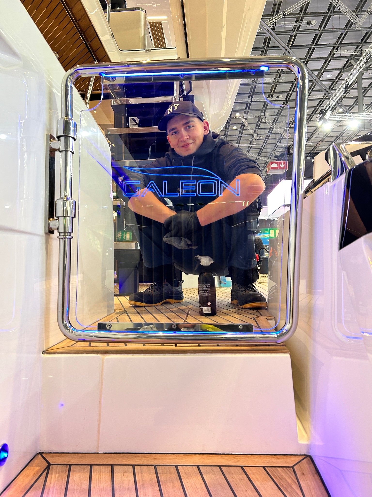 Mężczyzna w czapce NY i rękawicach, kuca w wejściu do kabiny jachtu, obok butelka środka czyszczącego, widoczna podświetlana nazwa 'GALEON'.