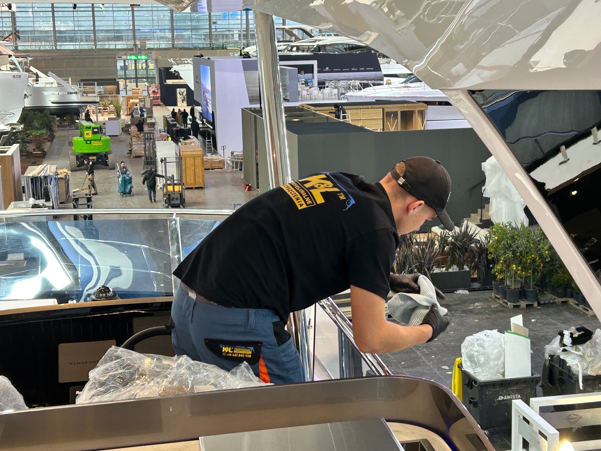 Pracownik w czarnej koszulce z logo firmy czyści wnętrze luksusowego jachtu na hali wystawienniczej, używając szmatki i rękawic ochronnych.