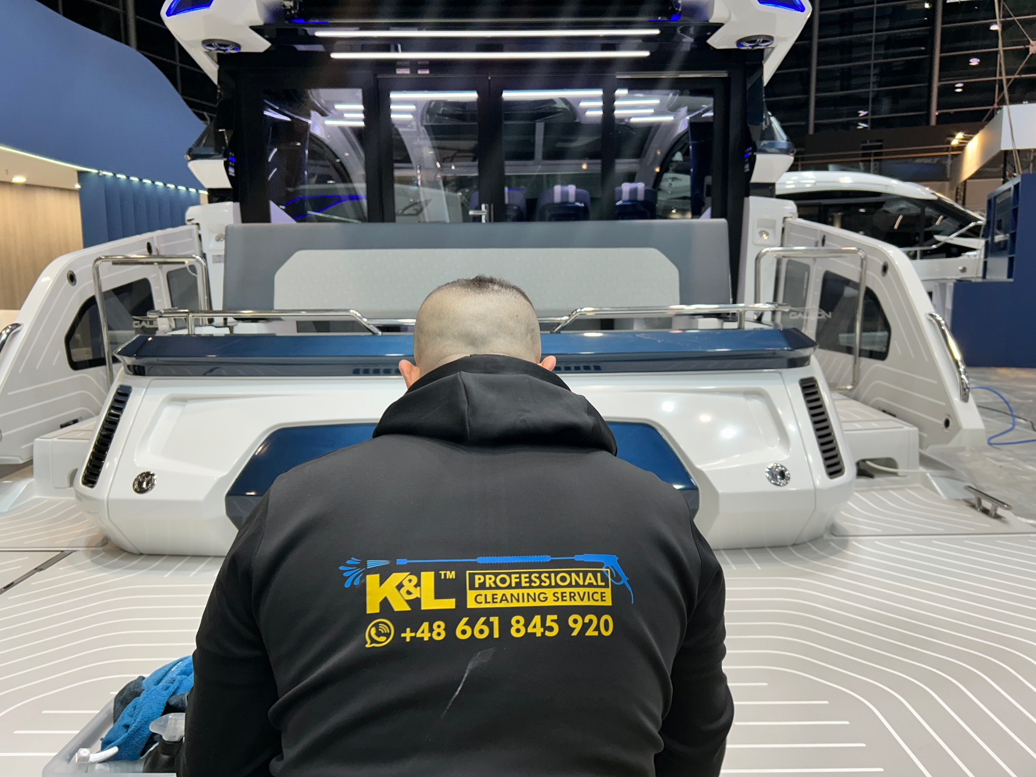 Osoba w bluzie z logo firmy czyszczącej, od tyłu, podczas pracy przy czyszczeniu wnętrza luksusowego jachtu na wystawie, widoczne detale wykończenia i akcesoria do czyszczenia.