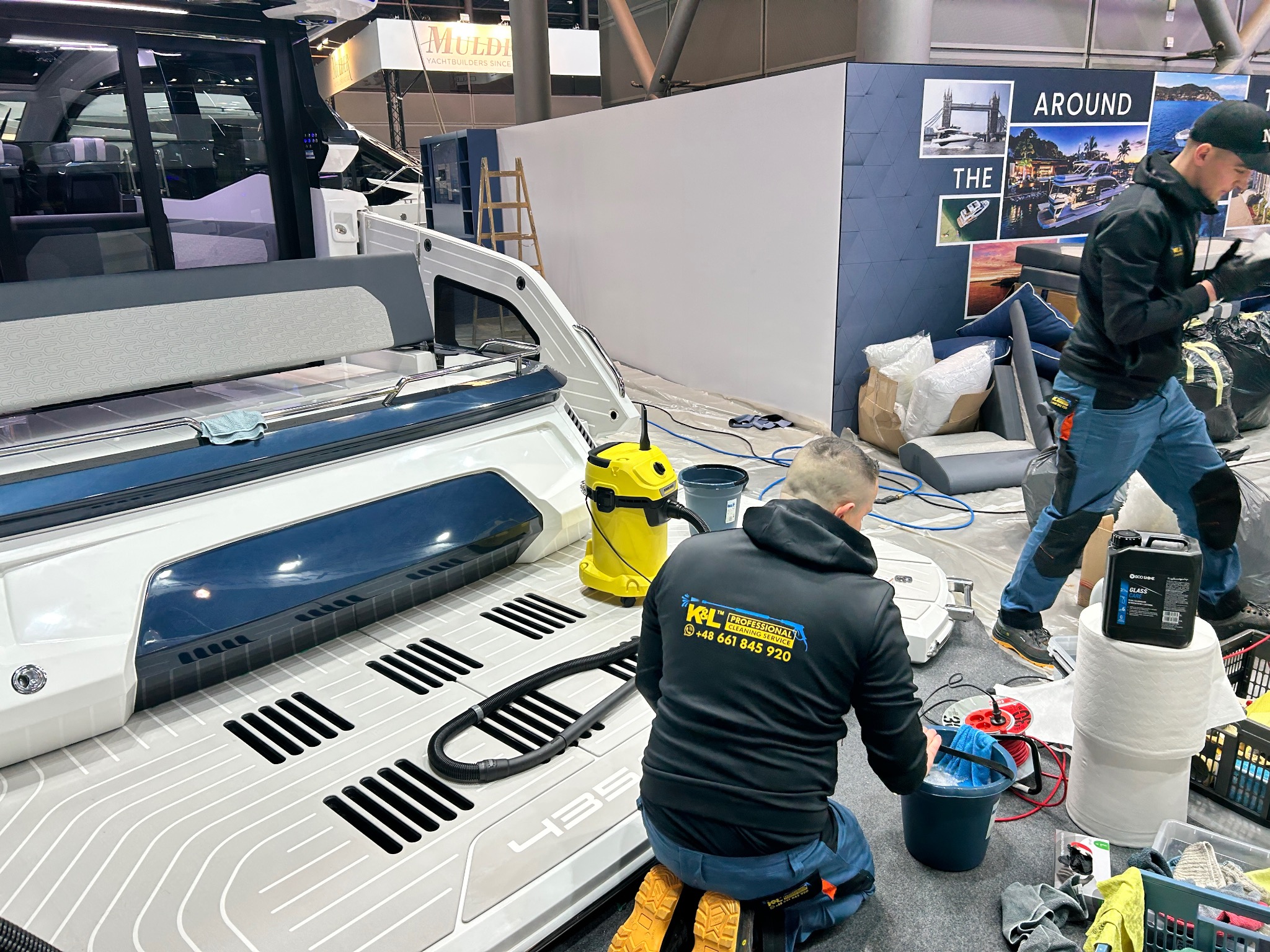 Dwaj mężczyźni w roboczych ubraniach czyszczą wnętrze luksusowej łodzi na wystawie, używając odkurzacza przemysłowego i środków czyszczących, widoczny żółty odkurzacz i butelka środka...