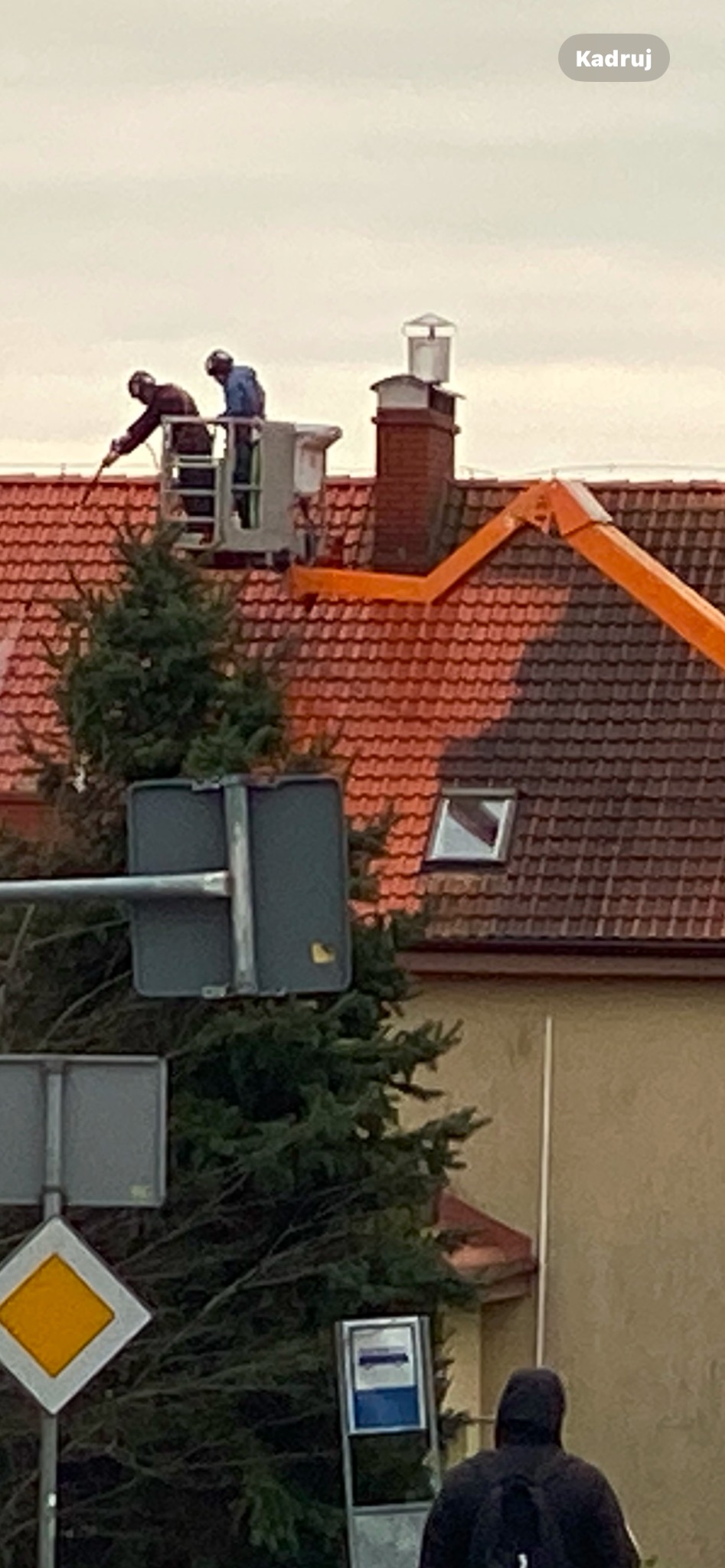 Pracownicy na podnośniku koszowym czyszczący dach z czerwonej dachówki, widoczny komin i okno dachowe, w tle drzewo i budynek, ujęcie z dołu.