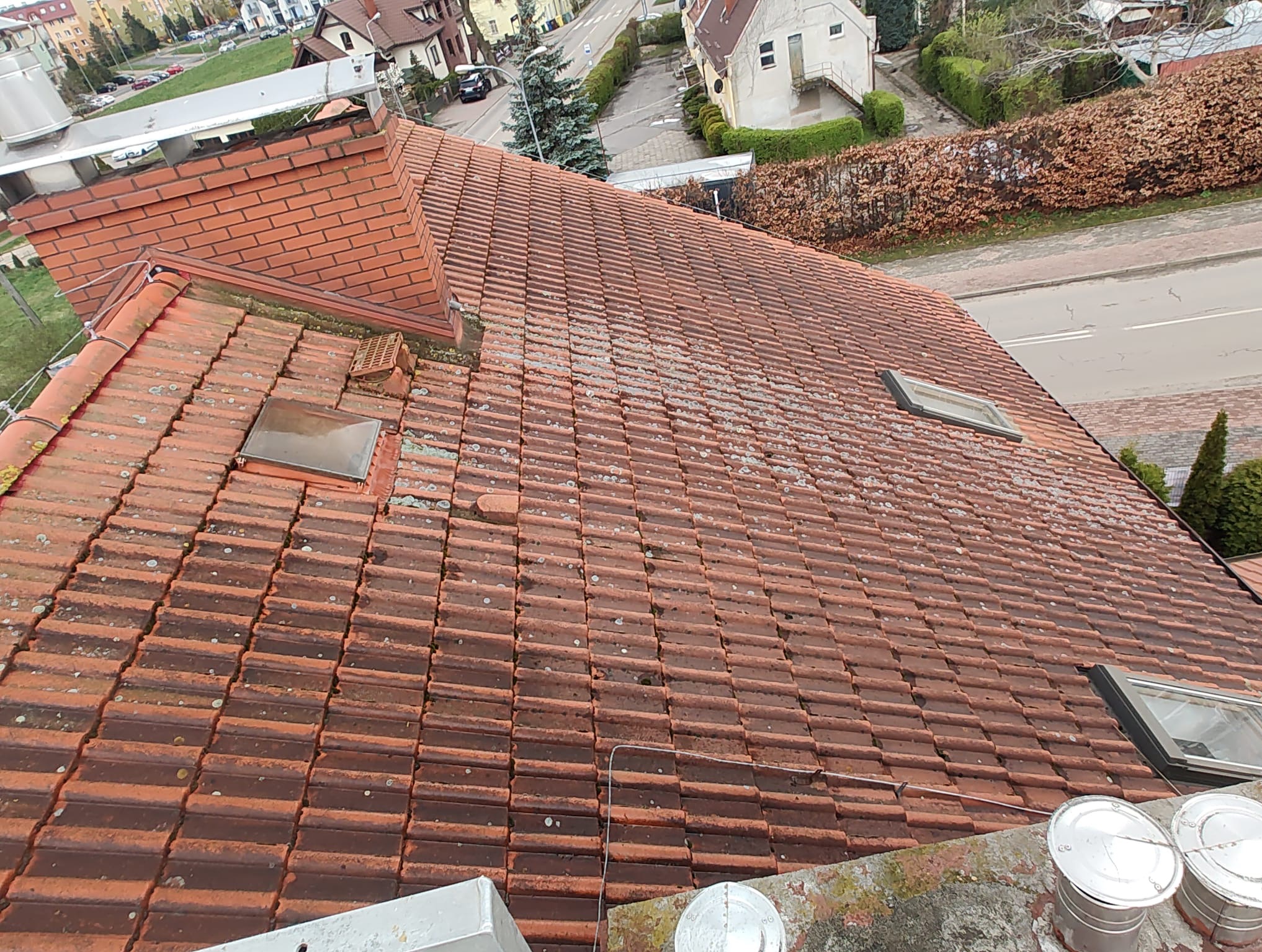 Czerwona dachówka z widocznymi śladami mchu i zabrudzeń, okna dachowe, komin z cegły, puszki po farbie w dolnym rogu, budynki mieszkalne w tle.