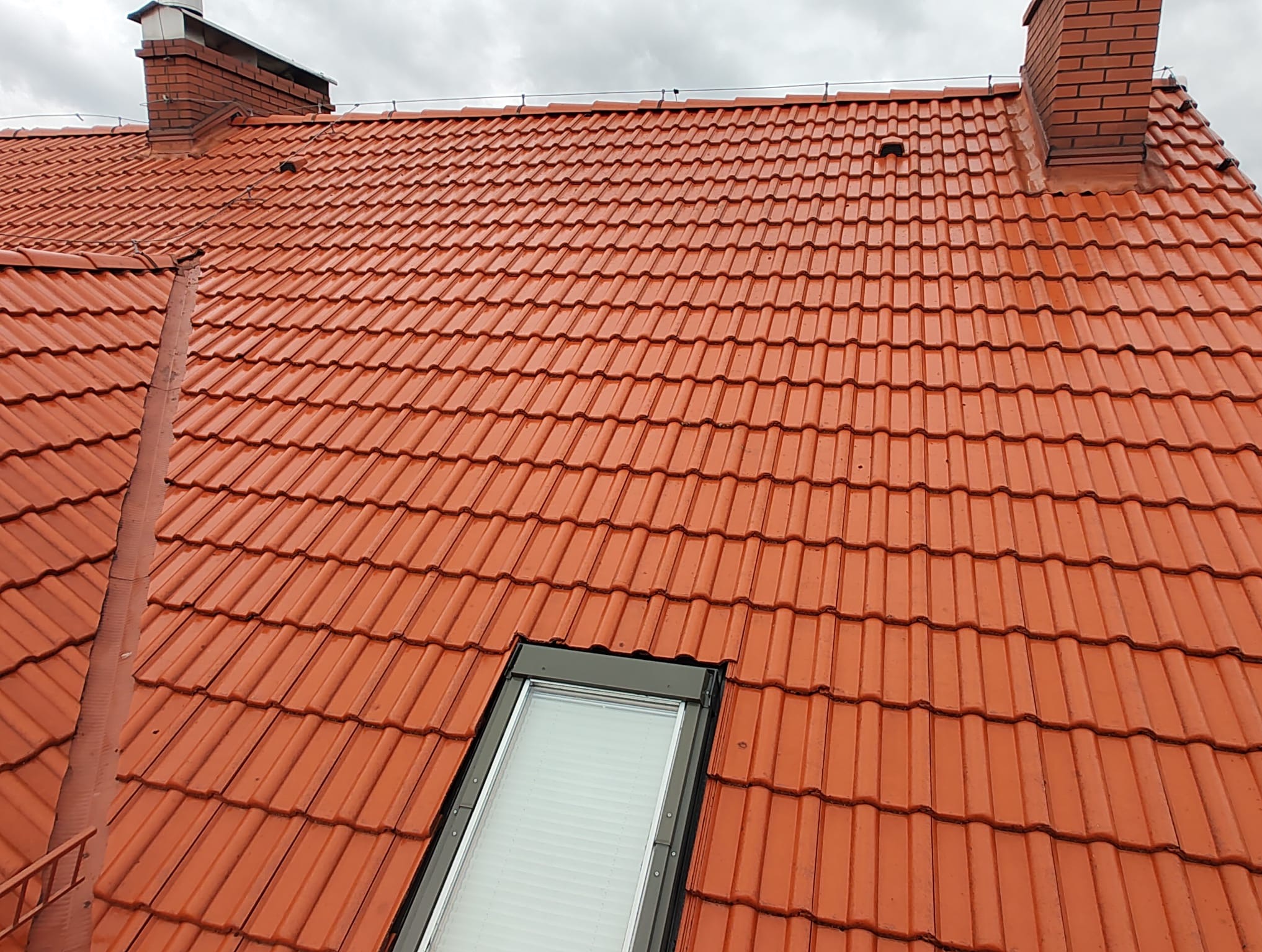 Czerwona dachówka ceramiczna z oknem dachowym i ceglanym kominem, widok z góry na pochmurnym tle.