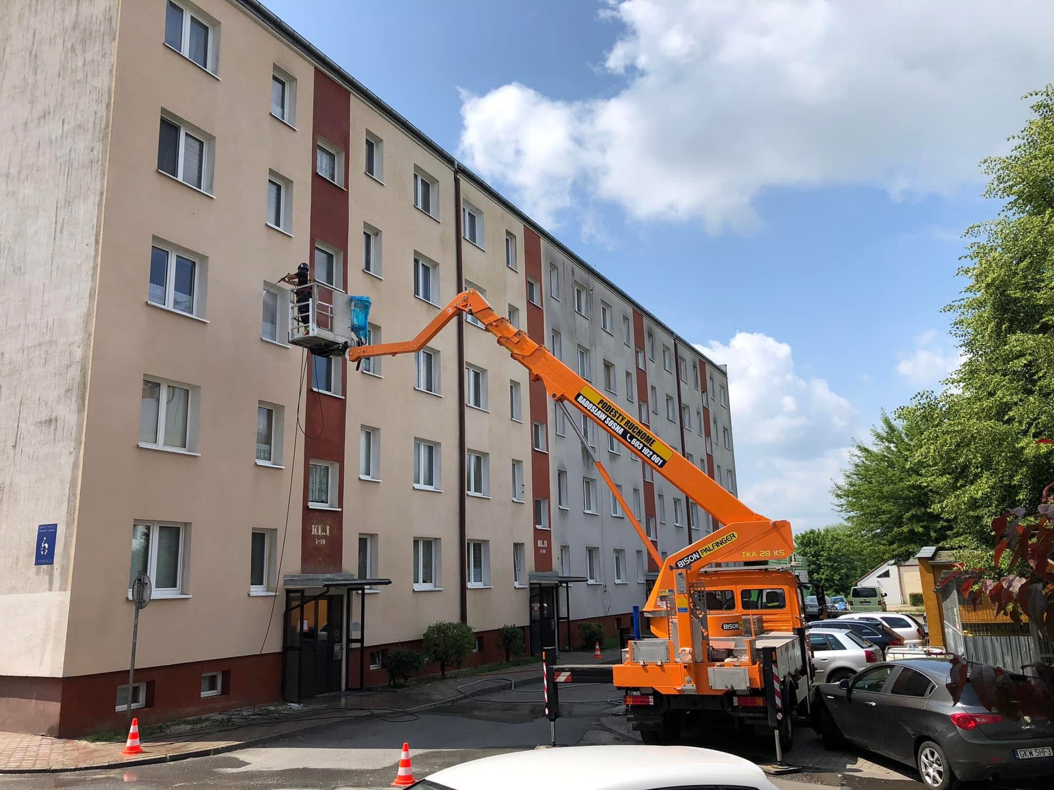 Pracownik na podnośniku koszowym czyści elewację budynku wielorodzinnego, widoczne ślady wody spływającej po ścianie, na pierwszym planie zaparkowane samochody i pachołek ostrzegawczy.