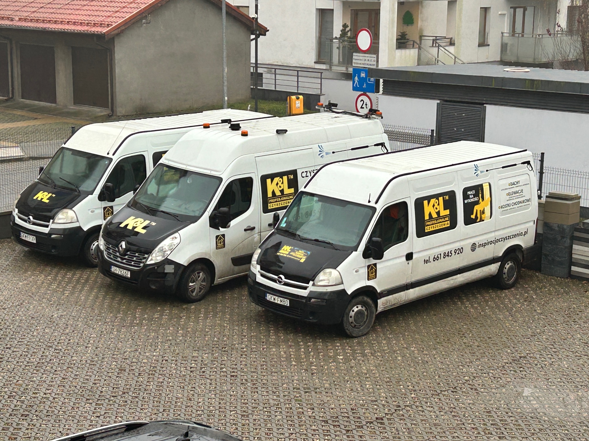 Trzy białe furgonetki firmy K&L zaparkowane na mokrej kostce brukowej, z widocznymi logo i numerami telefonów, w tle budynki mieszkalne i znaki drogowe.