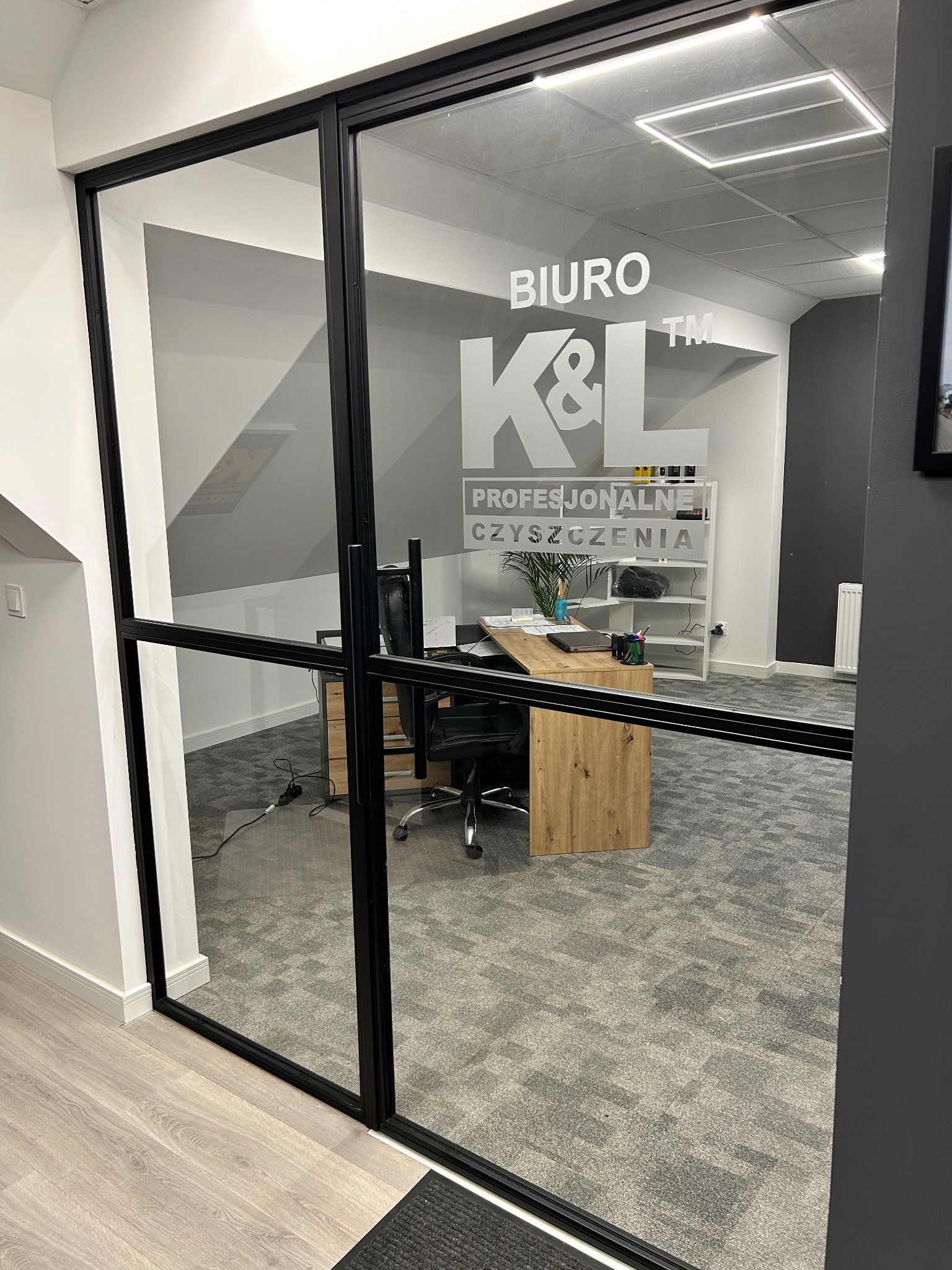 Szklane drzwi z napisem 'BIURO K&L PROFESJONALNE CZYSZCZENIA', widok na biuro z biurkiem, krzesłem i regałem.