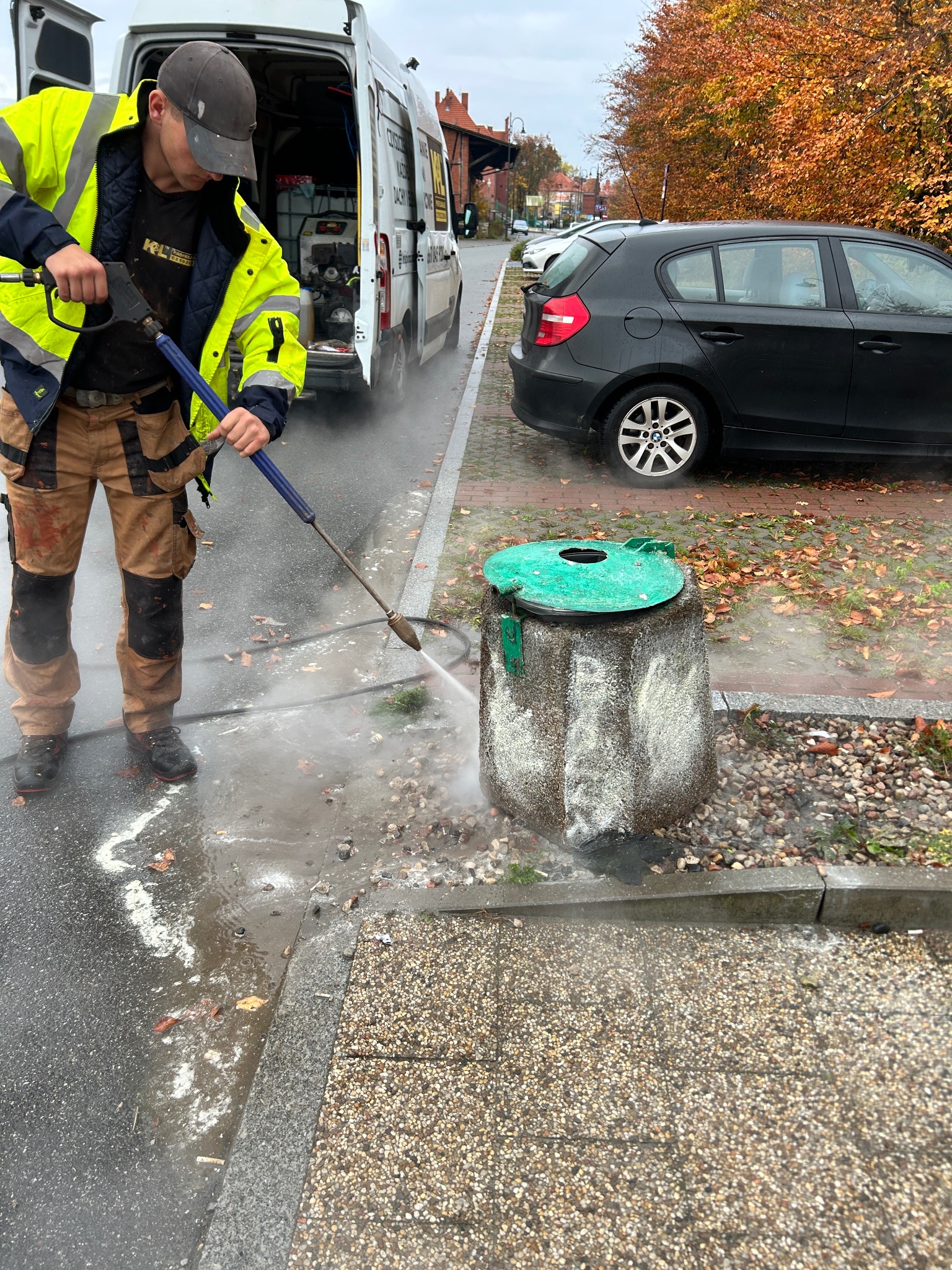 Pracownik w odblaskowej kurtce czyści ciśnieniowo betonowy kosz na śmieci z zieloną pokrywą na tle jesiennego krajobrazu miejskiego z zaparkowanym samochodem i furgonetką z wyposażeniem...