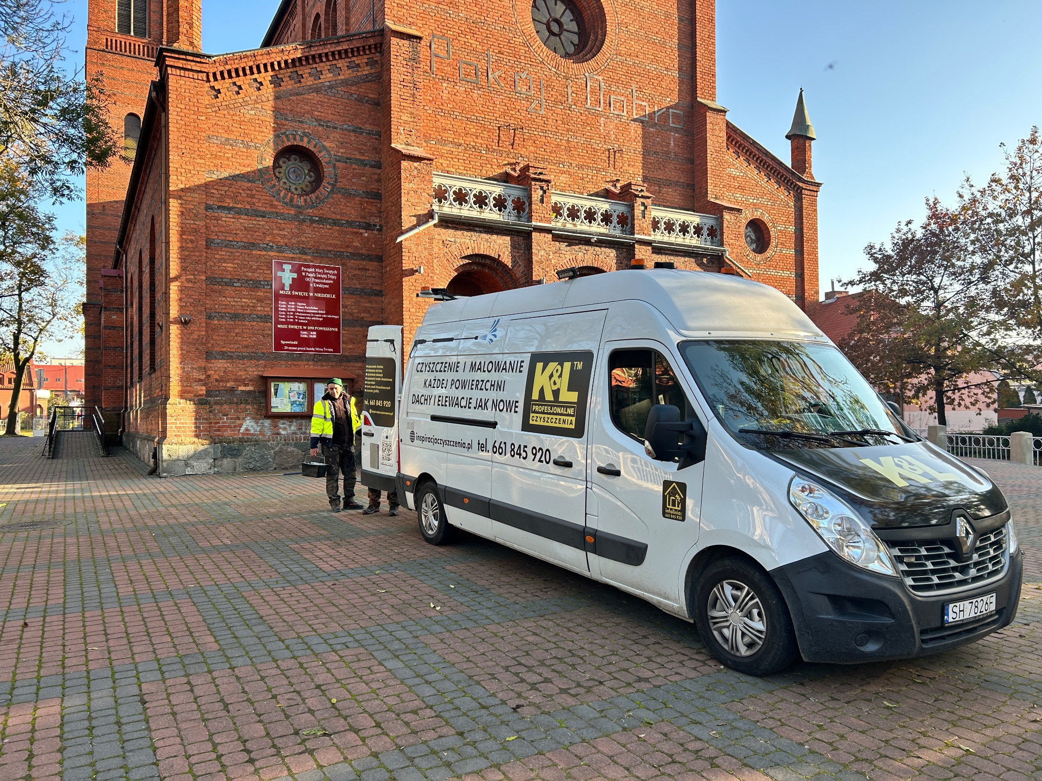 Ekipa czyszcząca z firmowym vanem K&L na tle ceglanego kościoła z tablicą informacyjną, kostka brukowa na pierwszym planie.
