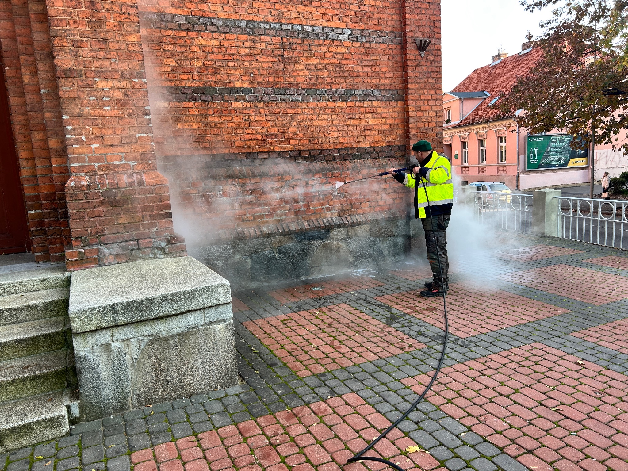 Pracownik w jaskrawej kamizelce odblaskowej czyści czerwoną ceglaną elewację budynku strumieniem wody pod ciśnieniem, widoczna para wodna i czysta powierzchnia, na pierwszym planie kostka brukowa.