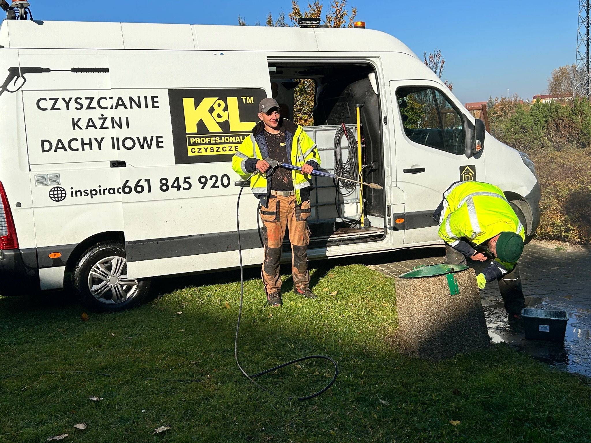 Dwóch pracowników w odblaskowych kurtkach czyści elewację kostki brukowej przy użyciu myjki ciśnieniowej i wiadra z wodą, na tle białego samochodu dostawczego z logo firmy i numerem telefonu.