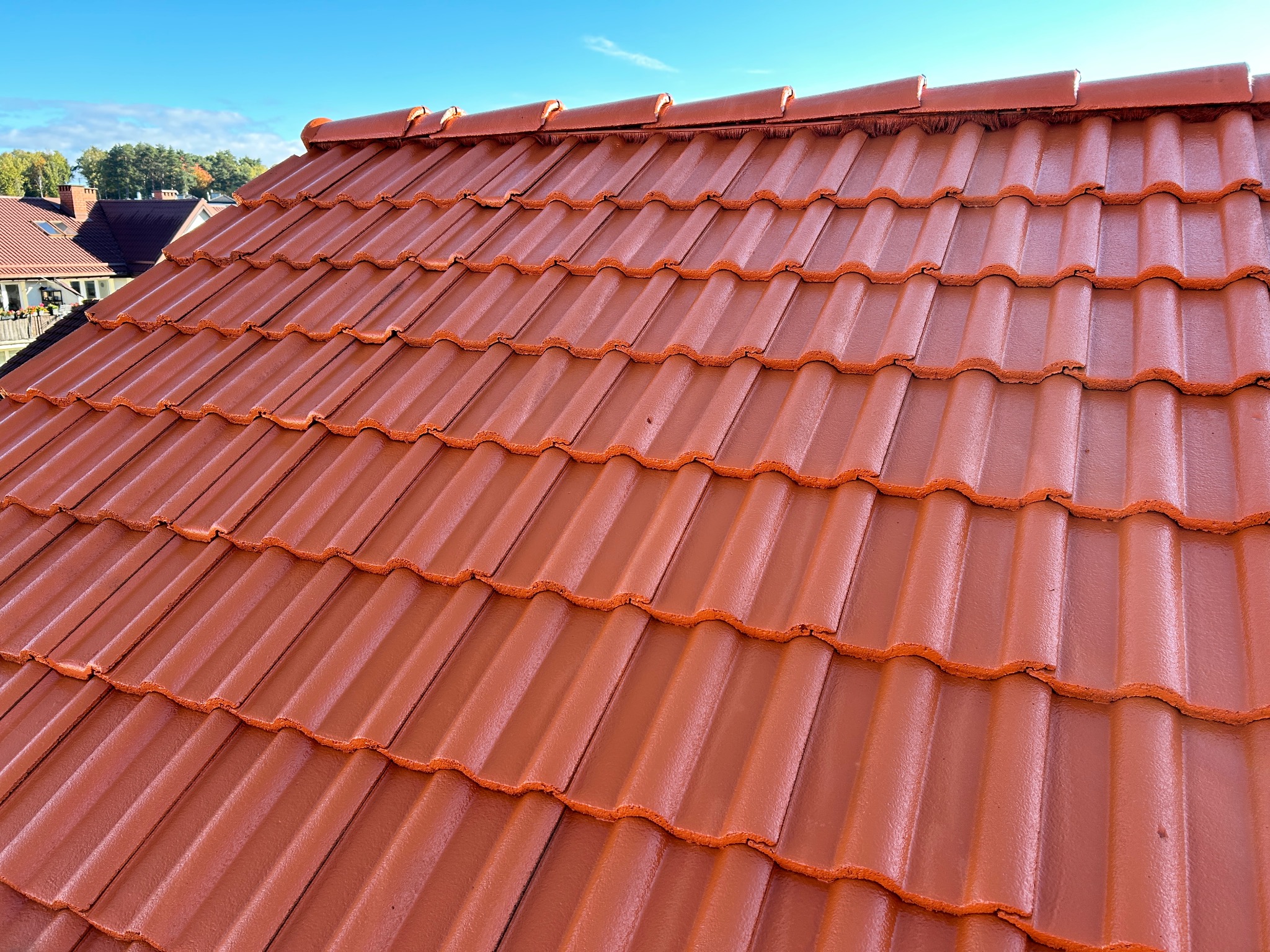 Błyszcząca, pomarańczowo-czerwona dachówka ceramiczna na dachu domu, widok z dołu pod kątem, fragment nieba w tle.