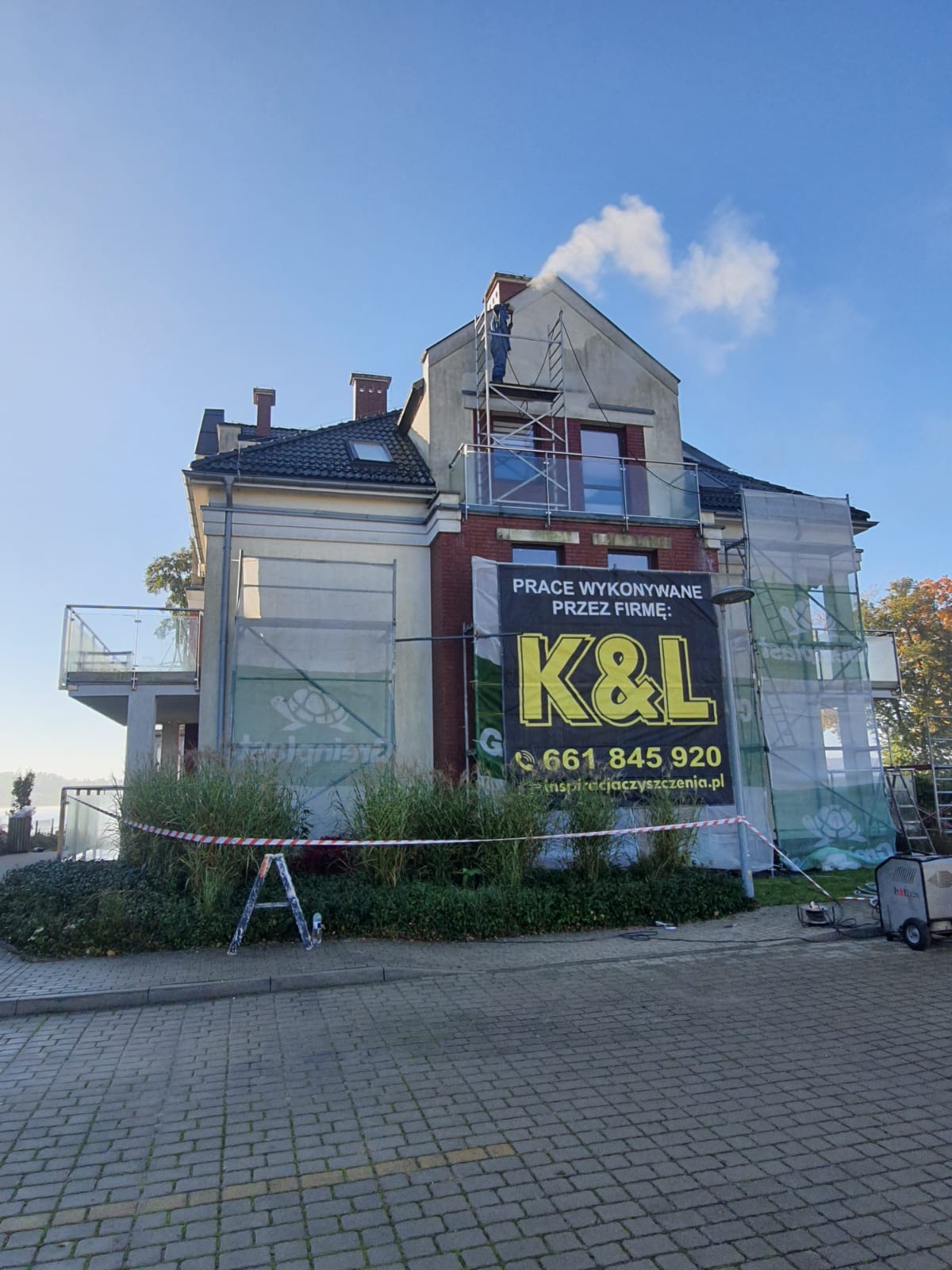 Pracownik na rusztowaniu czyści komin domu jednorodzinnego pod ciśnieniem, widoczny baner reklamowy firmy K&L oraz taśma ostrzegawcza.