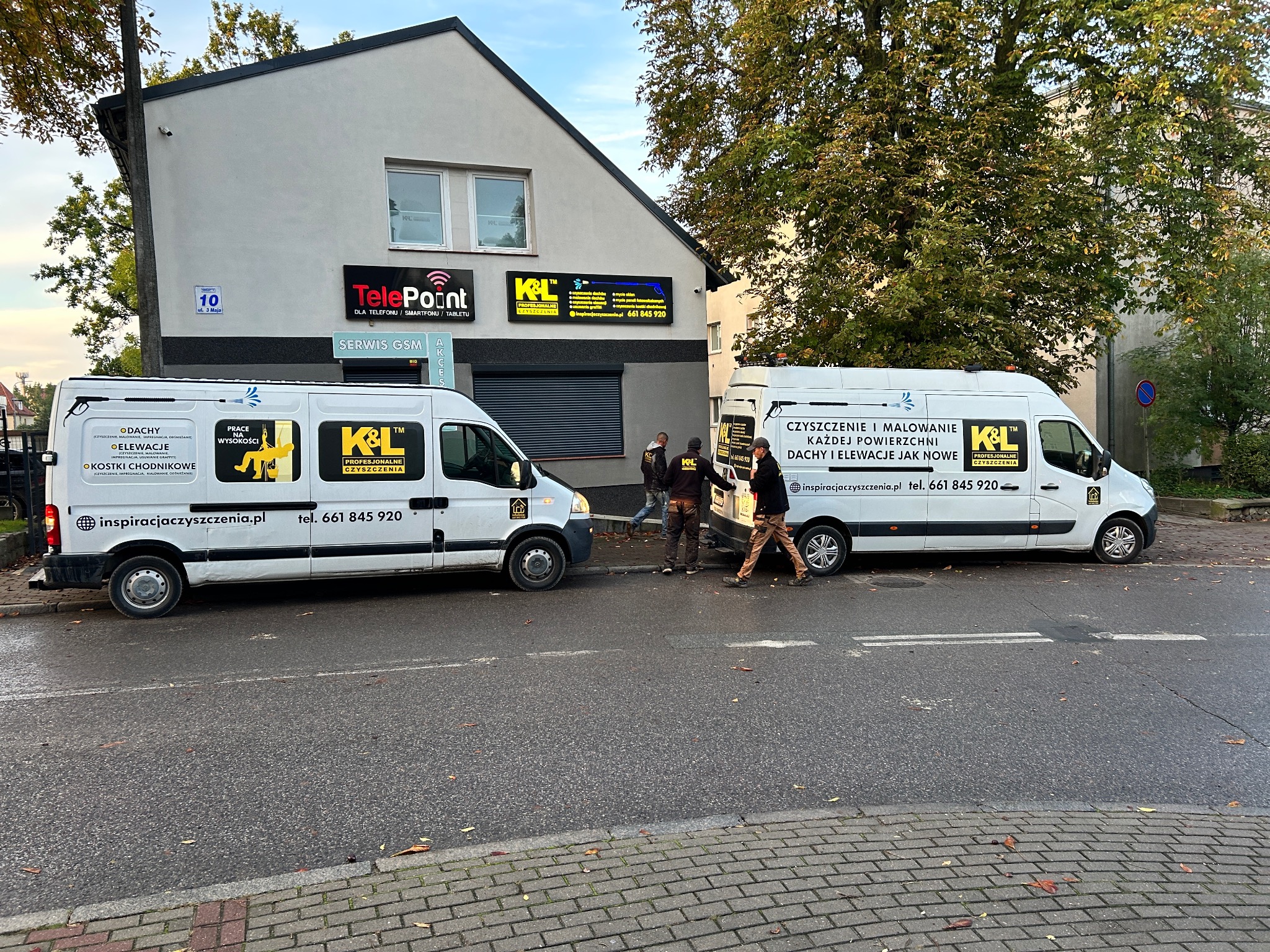 Dwa białe busy firmy czyszczącej z logo K&L zaparkowane przed budynkiem usługowym; pracownicy w ubraniach roboczych stoją między pojazdami.
