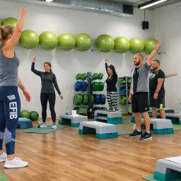 EFIB dr Magdalena Górecka - Grupowe zajęcia fitness w jasnej sali: kobiety i mężczyźni ćwiczą z podniesionymi rękami, zielone piłki i stepy w tle. Widok z perspektywy osoby ćwiczącej.