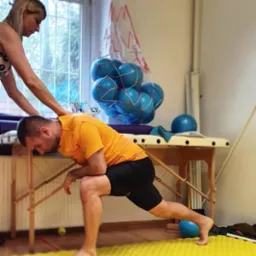 Mężczyzna w pomarańczowej koszulce wykonuje ćwiczenie rozciągające w obecności kobiety, która koryguje jego postawę; w tle widoczny stół do masażu i siatka z niebieskimi piłkami.